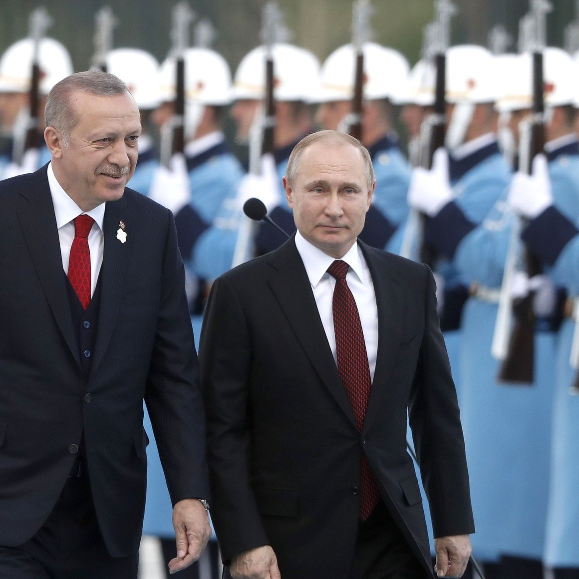 L'image montre deux dirigeants en train de marcher ensemble lors d'une cérémonie officielle. Ils sont entourés d'une garde d'honneur portant des uniformes distinctifs, avec des fusils à la main. Les deux hommes sont habillés en costumes formels, l'un ayant une cravate rouge et l'autre une cravate sombre. L'atmosphère semble solennelle, reflétant un événement diplomatique important.