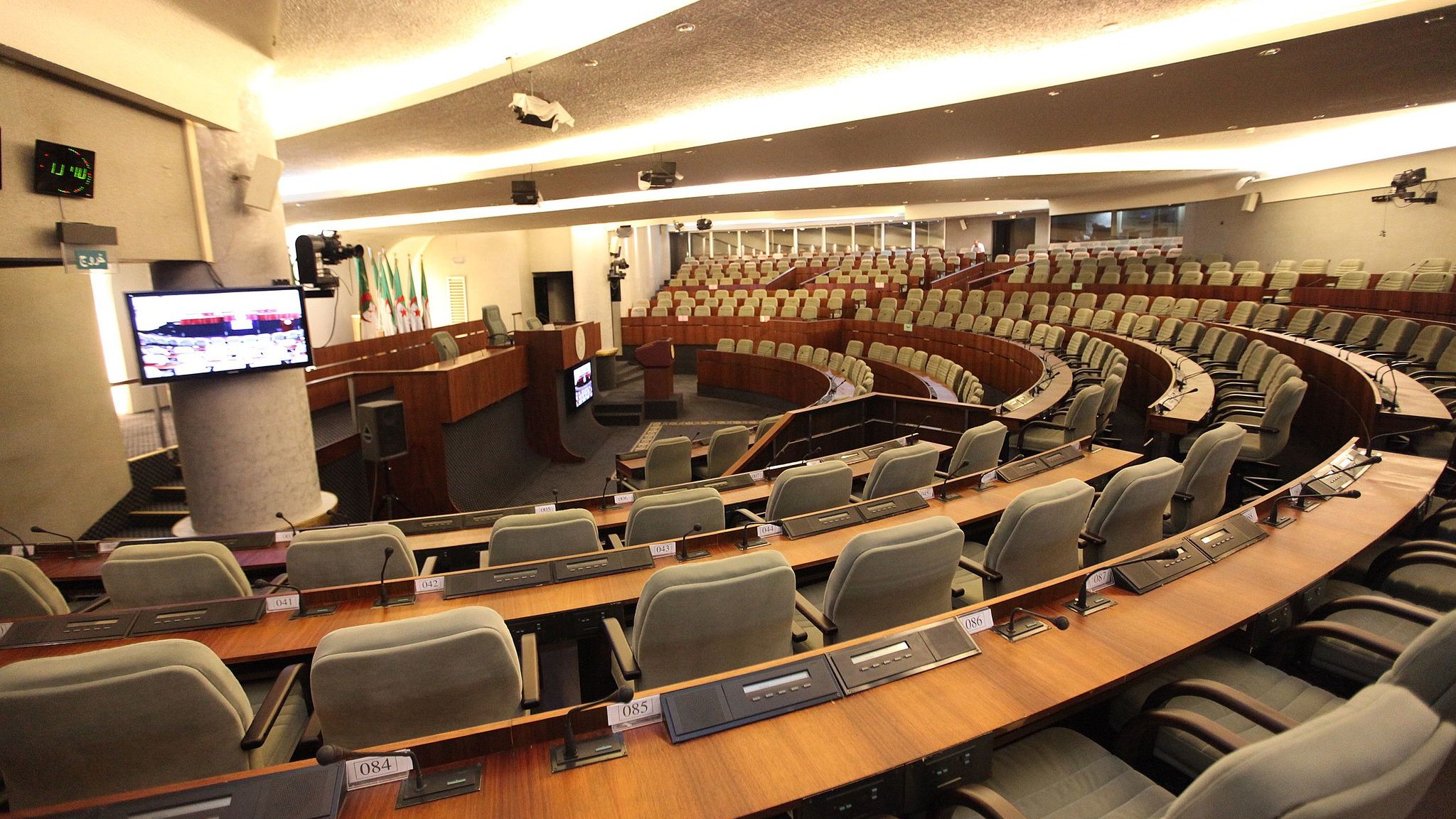 L'image montre une salle de réunion ou de débat, probablement une chambre législative. On y voit plusieurs rangées de sièges grisés disposés en arc de cercle autour d'une scène centrale. À l'avant, un bureau ou une tribune est visible, avec des équipements audiovisuels, comme un écran et des micros intégrés dans les tables. L'éclairage est doux et moderne, créant une atmosphère propice aux discussions. Les murs sont revêtus de matériaux contemporains, donnant une impression de sérieux et de fonctionnalité.
