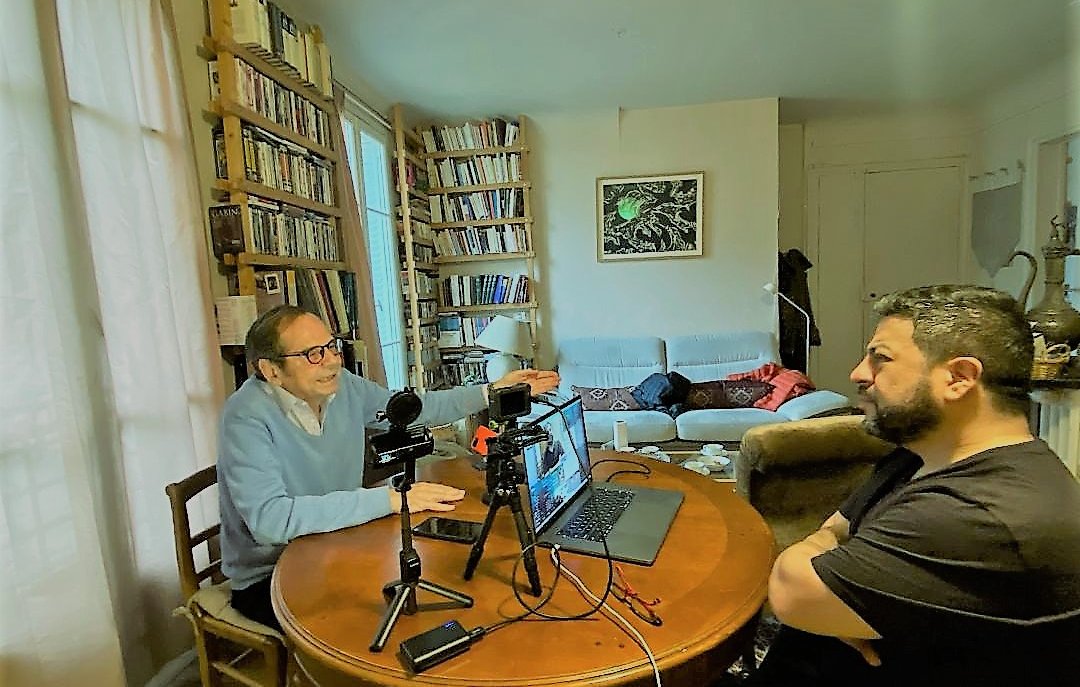 تظهر في الصورة مشهداً من داخل غرفة معيشة. على الطاولة الدائرية توجد أجهزة كمبيوتر وكاميرات، حيث يجلس شخصان يتحدثان معًا. الشخص الأول يرتدي سترة زرقاء ويبدو أنه يتحدث بفعالية، بينما الشخص الثاني يجلس مقابله، عابسًا أو متفكرًا. في الخلفية، توجد رفوف مليئة بالكتب، وأريكة مريحة، وظهر شخص آخر مستلقيًا على الأريكة. الإضاءة في المكان تبدو طبيعية، مما يعطي شعورًا بالراحة والتركيز على الحوار.