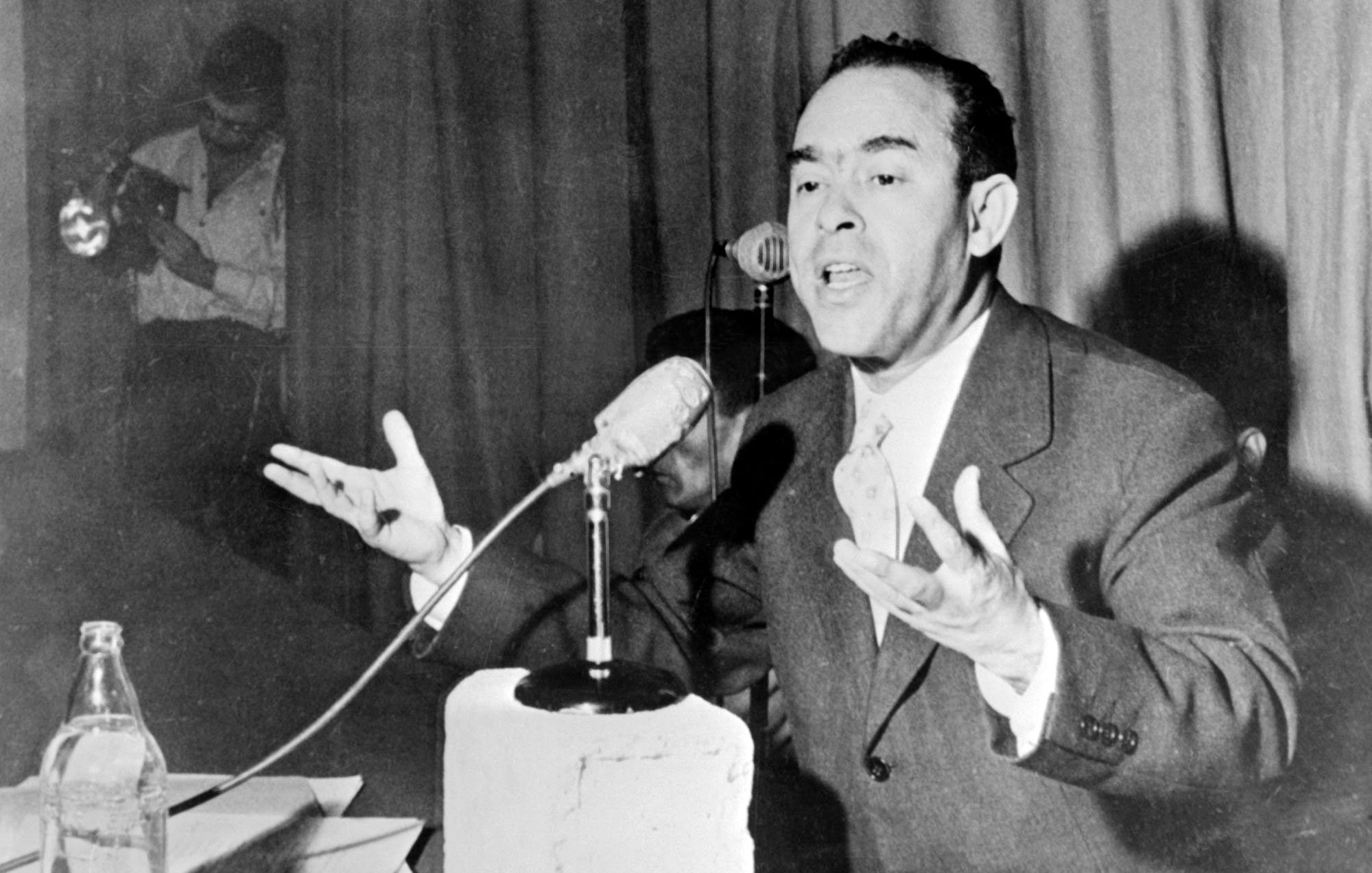 L'image montre un homme en train de s'exprimer avec enthousiasme lors d'une conférence ou d'un discours. Il est vêtu d'un costume et parle devant un microphone, gesticulant avec les mains pour accentuer ses propos. Sur la table devant lui, on peut voir un bloc de béton et quelques documents. En arrière-plan, une autre personne semble prendre des photos ou enregistrer. L'ambiance paraît sérieuse, soulignant l'importance de son discours.