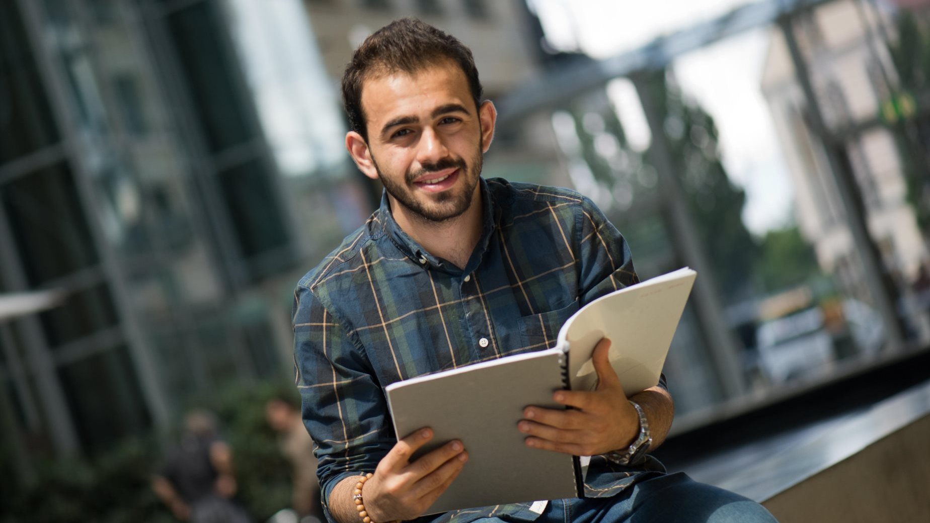 L'image montre un jeune homme assis sur un banc, souriant, tout en tenant un carnet ou un cahier dans ses mains. Il porte une chemise à carreaux et a l'air détendu, avec un environnement urbain en arrière-plan et des bâtiments modernes. La lumière naturelle éclaire la scène, créant une atmosphère agréable.