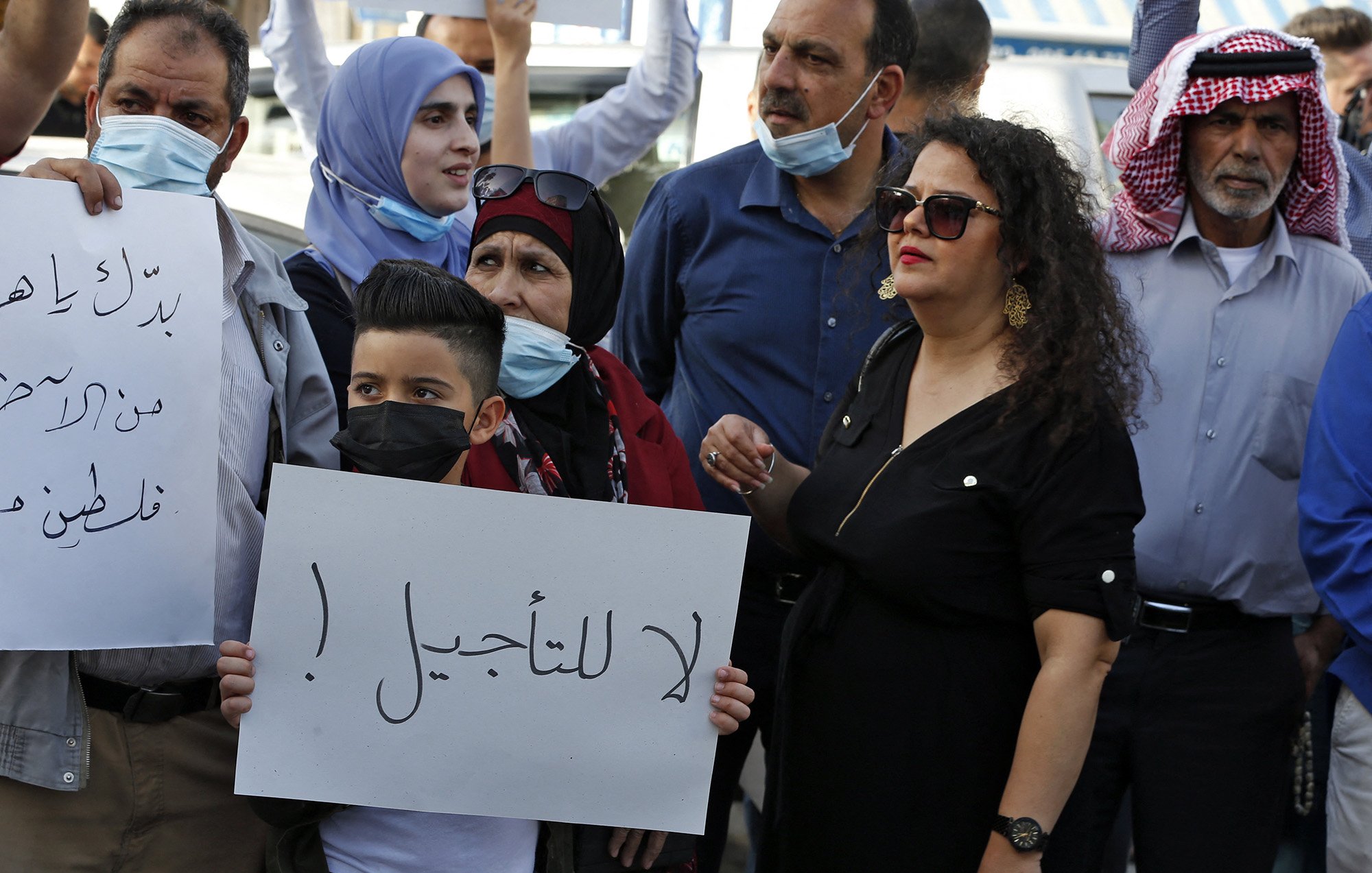 تظهر في الصورة مجموعة من الناس في مظاهرة. يرفع بعضهم لافتات، بينما يرتدي الآخرون كمامات. في المقدمة، يوجد طفل صغير يحمل لافتة مكتوب عليها "لا للتأجيل!"، وتحيط به مجموعة من البالغين، بينهم امرأة ترتدي فستانًا أسود وبعض الرجال بملابس تقليدية. تعكس الصورة جوًا من الاحتجاج والتعبير عن الرأي.