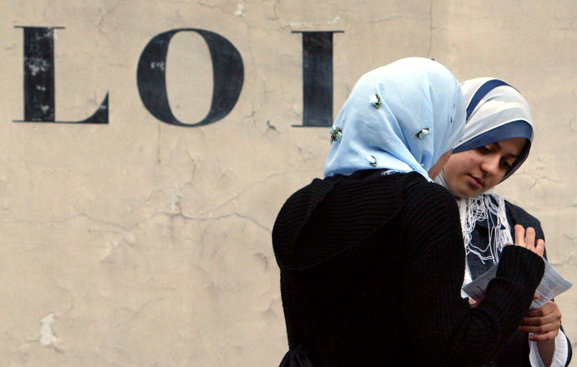 L'image présente deux femmes discutant, l'une d'elles portant un hijab bleu clair et l'autre un hijab sombre. Elles semblent concentrées sur un document ou une feuille qu'elles regardent ensemble. En arrière-plan, on peut voir le mot "LOI" écrit en lettres majuscules sur un mur beige, ce qui donne un contexte à la scène. L'atmosphère semble calme et introspective.