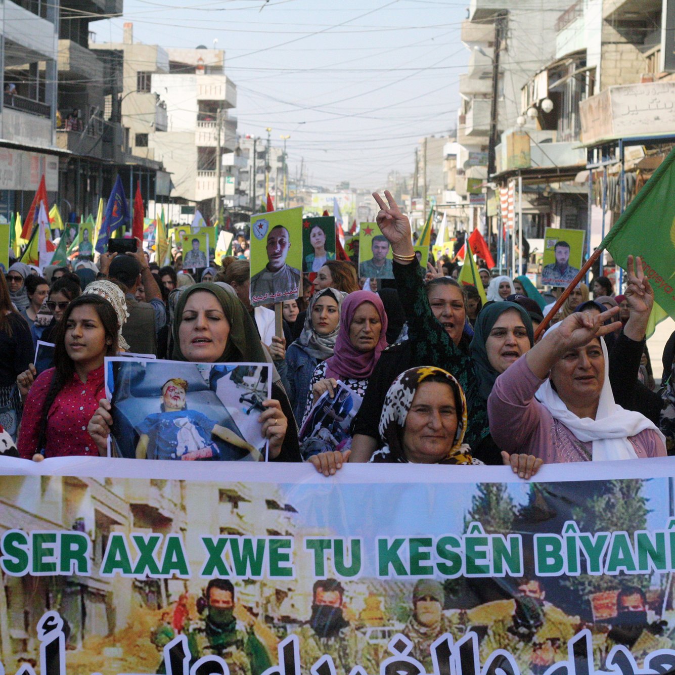 L'image montre une grande foule de personnes, principalement des femmes, participant à une manifestation. Elles portent des drapeaux colorés et affichent des pancartes avec des slogans. Les manifestants semblent exprimer des revendications et un soutien, probablement liées à des questions politiques ou sociales. L'ambiance est dynamique et déterminée, avec des signes de solidarité parmi les participants. Les rues environnantes sont visibles, ainsi que des bâtiments en arrière-plan.