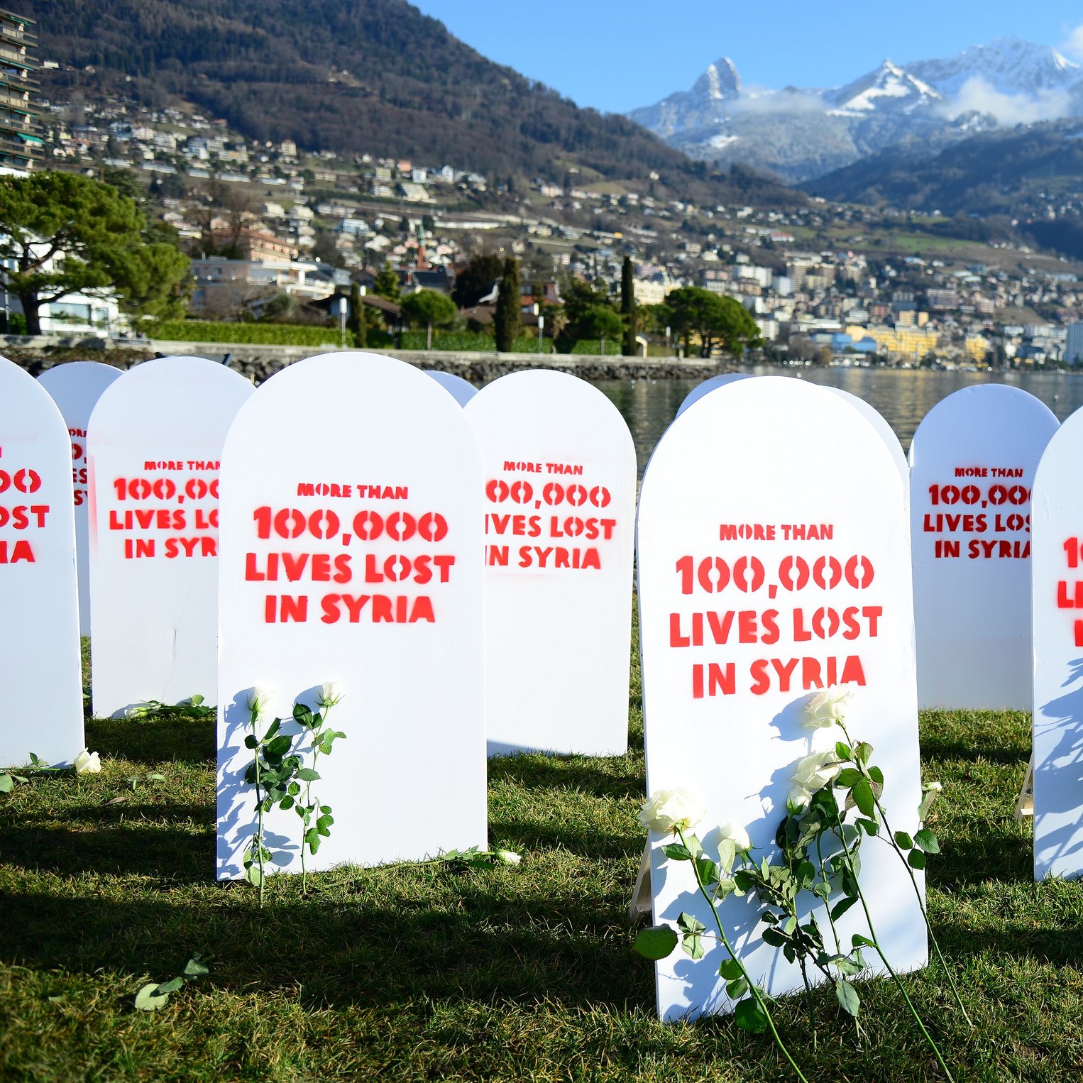 L'image montre un rassemblement commémoratif avec plusieurs pierres tombales blanches disposées sur un terrain vert. Chaque pierre tombale est marquée de rouge avec des inscriptions telles que "More than 100,000 lives lost in Syria". À côté des pierres tombales, il y a des roses ou des plantes, symbolisant probablement la mémoire des vies perdues. En arrière-plan, on peut voir une vue magnifique de montagnes et d'une ville au bord de l'eau. L'ambiance générale semble être solennelle et réfléchissante, soulignant une tragédie humanitaire.