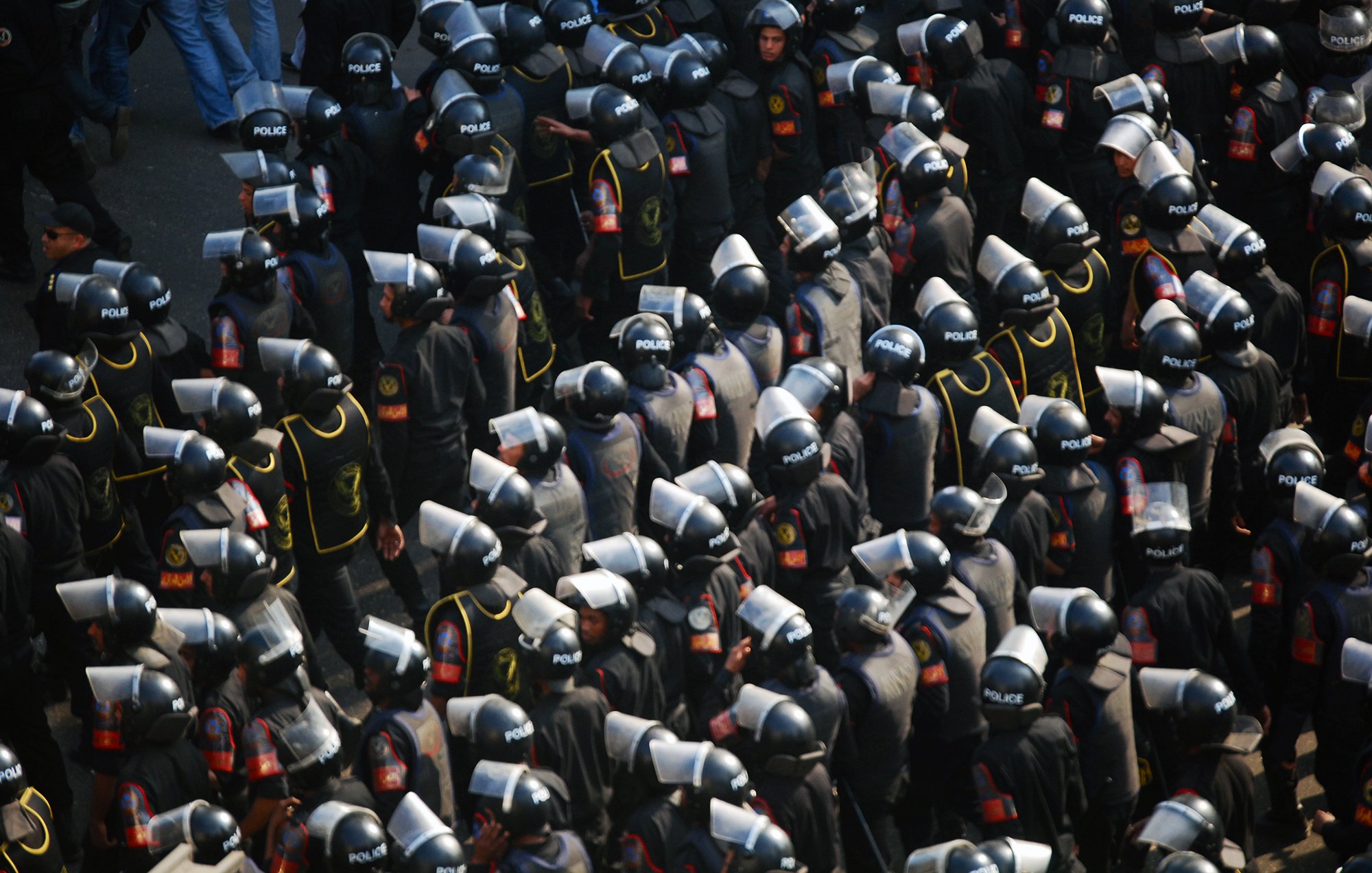 L'image montre un grand groupe de policiers en tenue d'émeute. Ils portent des casques et des boucliers, et sont regroupés de manière organisée. Les uniformes sont sombres, avec des détails visibles tels que des insignes ou des logos indiquant leur affiliation. L'ensemble donne une impression de préparation et de contrôle, évoquant un contexte de maintien de l'ordre lors d'une situation potentiellement tendue.