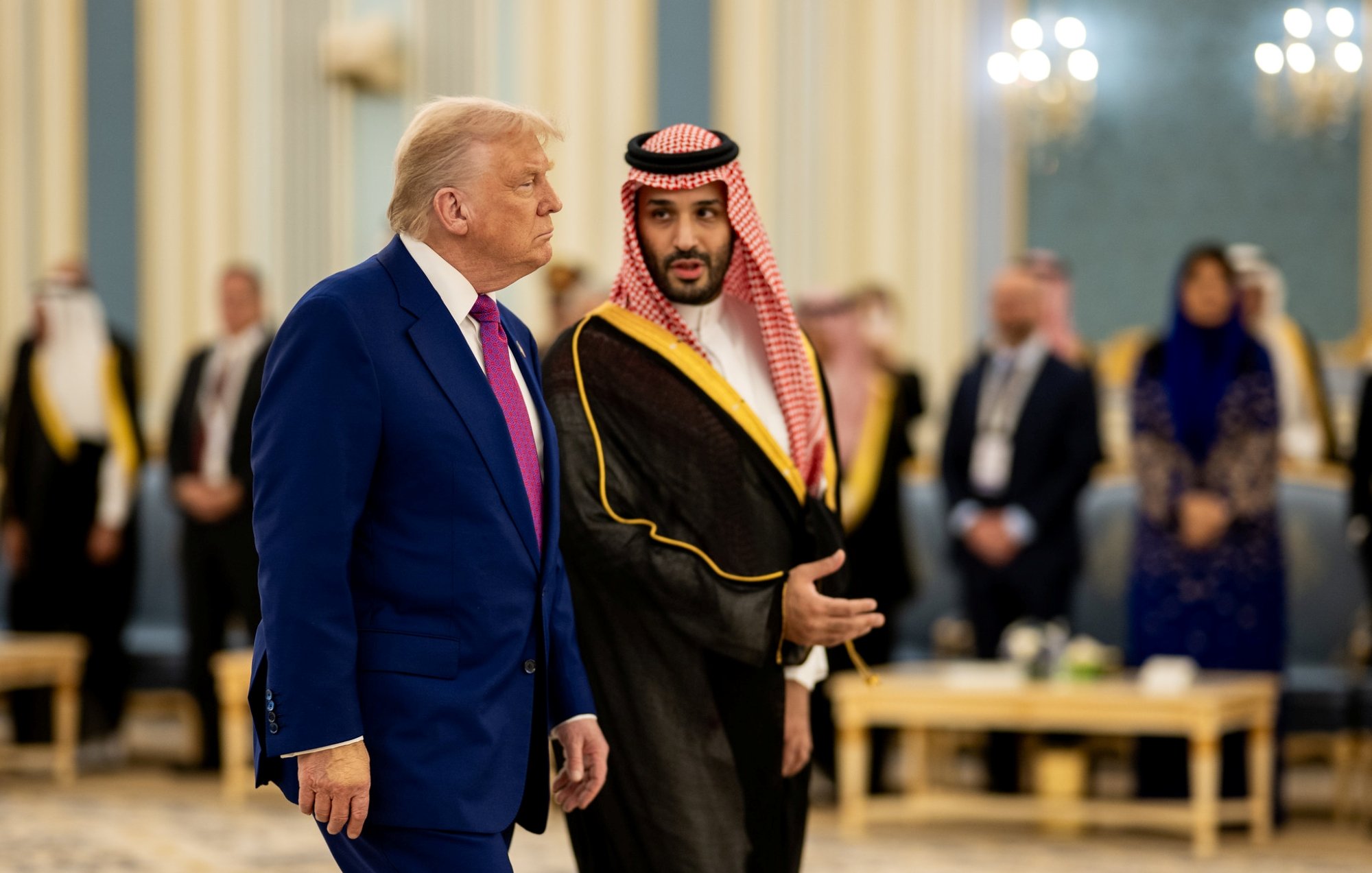 دو مرد در لباس‌های رسمی مختلف در یک محیط مجلل با نور ملایم و حضور افرادی دیگر.