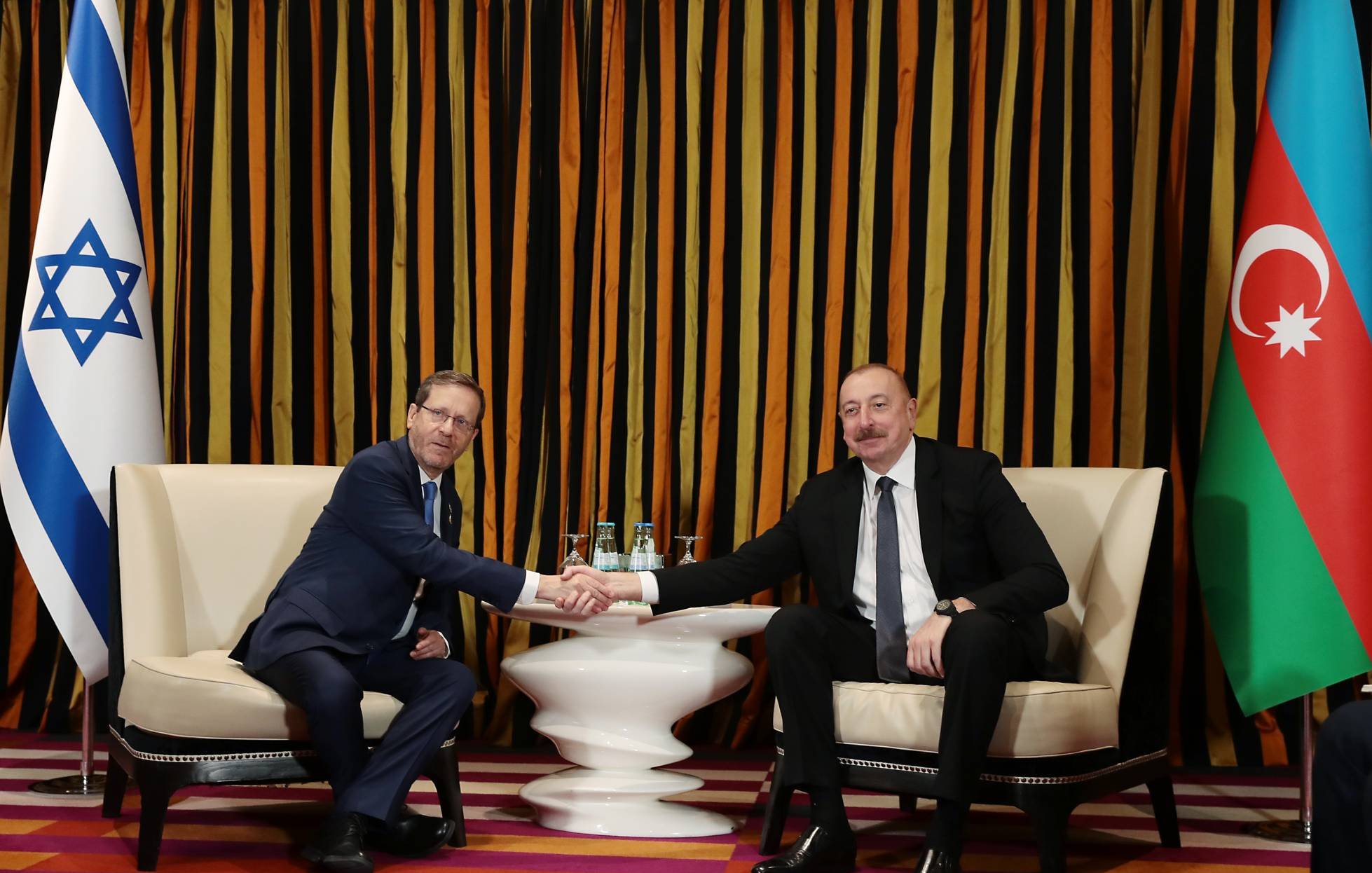 دو مرد در حال دست دادن نشسته‌اند. پرچم‌های اسرائیل و آذربایجان در پس‌زمینه هستند. رنگ‌ها گرم و زنده‌اند.