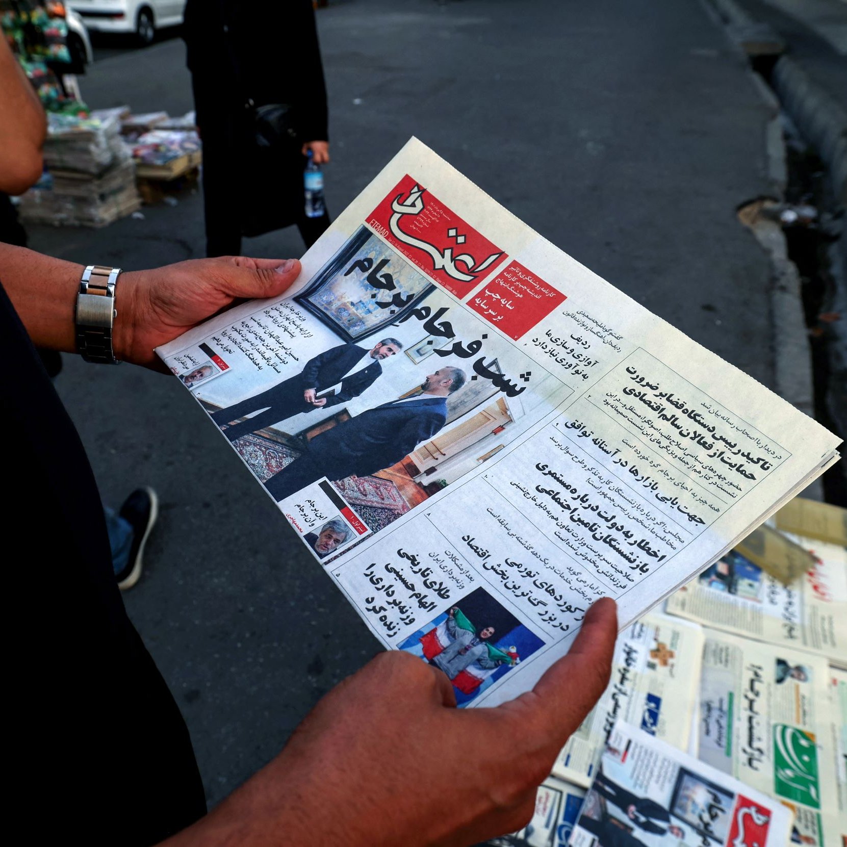 في الصورة، يُظهر شخص يحمل صحيفة باللغة العربية. تبدو الصحيفة جديدة ومطوية، ويظهر عنوان بارز على الصفحه الأولى، كما تُلاحظ مجموعة من الصحف في الخلفية. يبدو أن المشهد يدور في مكان عام، ربما في الشارع، حيث توجد بعض السيارات والأشخاص في محيط الصورة.