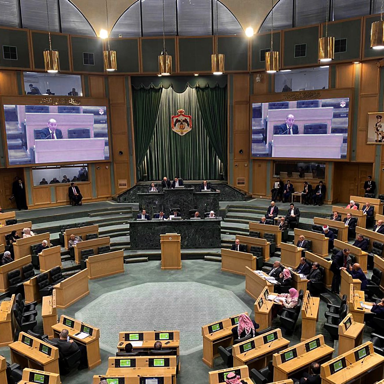 الصورة تظهر قاعة مجلس النواب، حيث يتم عقد جلسة. القاعة مزينة بخلفية خضراء ورموز وطنية، وتحتوي على شاشات عرض كبيرة. يتواجد عدد من النواب الذين يجلسون في مقاعدهم، بينما يظهر بعضهم يتحدث أو يستمع للخطاب الملقاة. الأجواء تعكس نشاطًا رسميًا يتمثل في النقاشات السياسية.