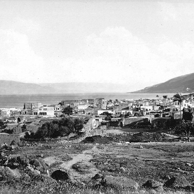 L'image montre un paysage historique d'un village côtier, probablement situé près d'un lac ou d'une mer, avec des bâtiments disposés le long de la promenade. Il y a un minaret ou une tour surmontée d'un dôme, indiquant la présence d'une culture musulmane. En arrière-plan, on peut voir des collines qui encadrent le paysage. Le ciel est légèrement nuageux, créant une ambiance tranquille. L'ensemble de l'image a un aspect noir et blanc, ce qui lui donne un caractère ancien.