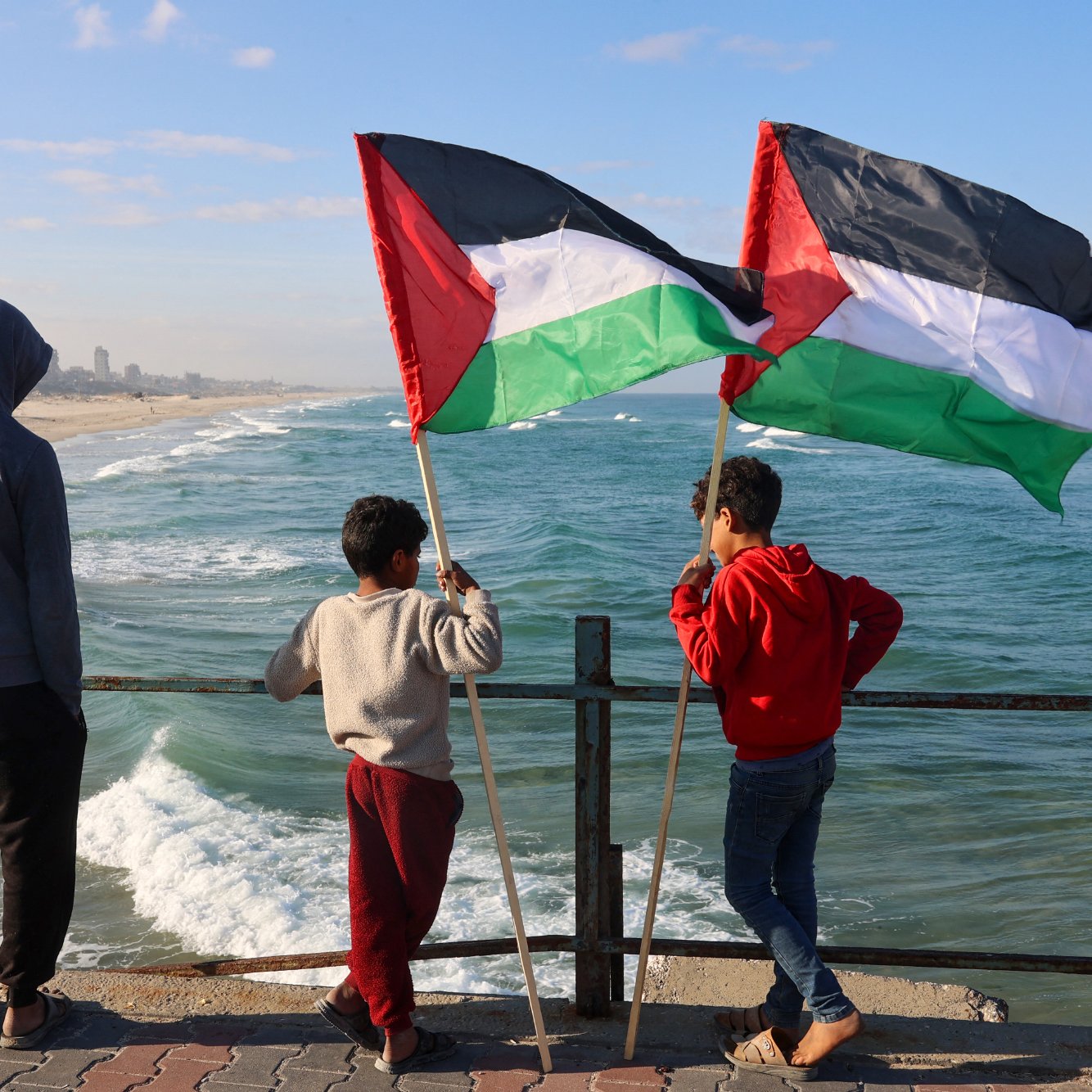 L'image montre trois enfants debout sur un bord de mer, regardant vers l'horizon. Deux des enfants tiennent des drapeaux avec les couleurs noir, rouge, vert et blanc, emblématiques de la Palestine. La mer est agitée, avec des vagues qui s'écrasent contre le rivage, et en arrière-plan, on peut apercevoir des bâtiments sur la plage. Les enfants semblent contemplatifs, profitant de l'instant. Le ciel est partiellement nuageux, créant une atmosphère tranquille.