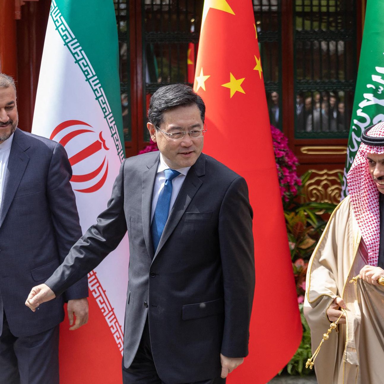 L'image montre trois hommes en costume, se tenant côte à côte devant des drapeaux. On devine que ces hommes représentent respectivement l'Iran, la Chine et l'Arabie Saoudite, compte tenu des drapeaux en arrière-plan. Ils semblent être en discussion lors d'un événement officiel. La scène est colorée, avec des éléments décoratifs comprenant des fleurs. Les hommes affichent des expressions sérieuses et engagées.