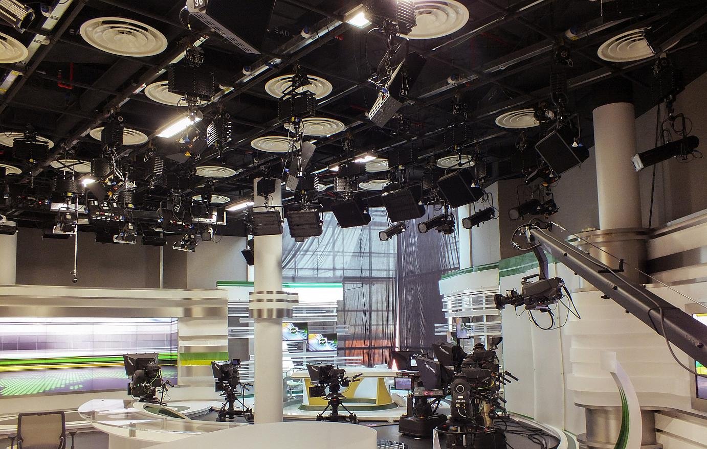 L'image montre un studio de télévision. On peut voir plusieurs caméras montées sur des tréteaux, prêtes à capturer des scènes. Le décor est moderne, avec des surfaces blanches et des accents de couleurs vives, notamment du vert. Des lumières suspendues au plafond éclairent le plateau, et il y a des écrans en arrière-plan, probablement utilisés pour afficher des informations ou des graphiques. L'ambiance semble professionnelle et organisée, typique d'un environnement de diffusion.