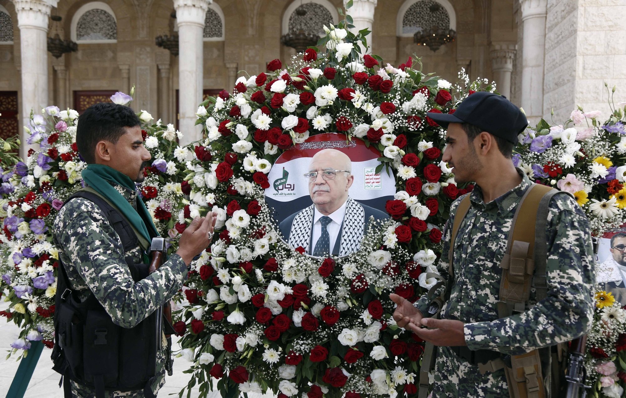 Deux hommes en uniforme discutent devant une couronne de fleurs avec un portrait au centre.