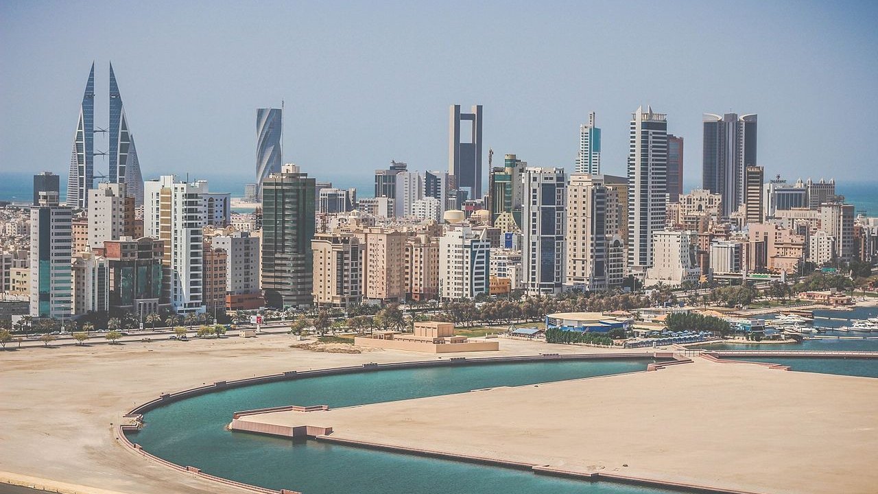 تظهر الصورة مشهداً لمدينة عصرية تضم العديد من ناطحات السحاب والمباني الحديثة. يتواجد في الخلفية مجموعة من الأبراج المميزة، بعضها ذو تصميم فريد. كما يظهر جزء من البحر على اليمين، مع مرفأ صغير يضم قوارب. السماء تبدو صافية وتضيء بأشعة الشمس، مما يضفي شعوراً بالوضوح والنشاط على المشهد العام.