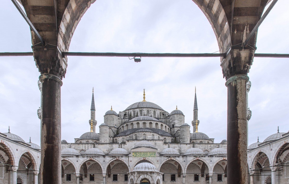 الصورة تظهر مشهدًا جميلًا لمدخل جامع السلطان أحمد في إسطنبول، المعروف أيضًا بالجامع الأزرق. يتوسط الصورة قبة كبيرة محاطة بأربعة مآذن طويلة. الأروقة المظللة تتواجد في المقدمة، حيث يوجد عدد من الأشخاص يتجولون ويتفاعلون مع المكان. السماء مغطاة بسحاب خفيف، مما يضفي جوًا مميزًا على المشهد. تصميم المعمار يجمع بين الأساليب الإسلامية التقليدية واللمسات الفنية الجميلة.