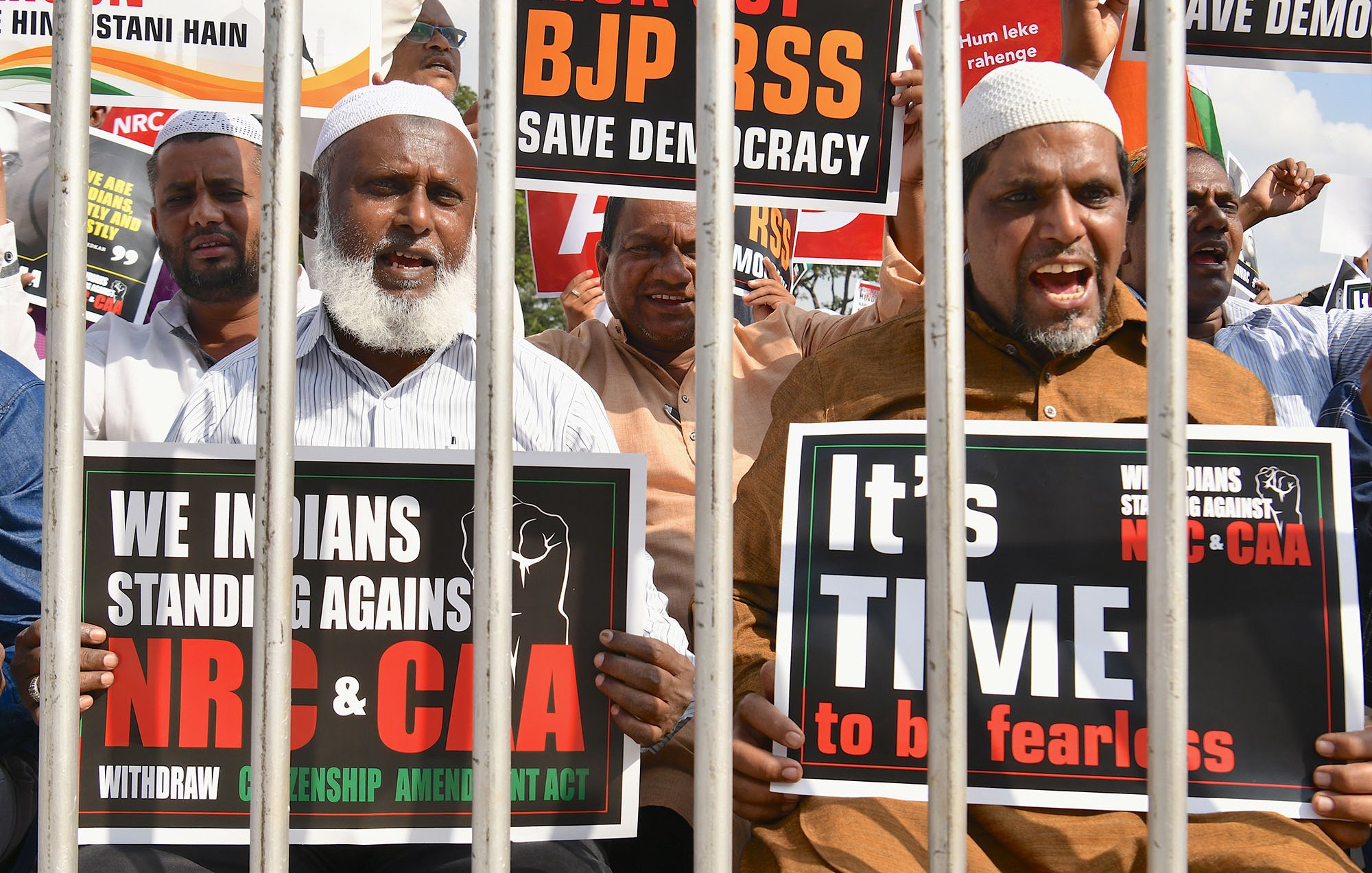 L'image montre un groupe de manifestants qui se tiennent derrière des barreaux. Ils tiennent des pancartes exprimant leur opposition à certaines lois et politiques en Inde, notamment le BJP, le NRC et le CAA. Les manifestants ont des expressions déterminées et sont clairement engagés dans leur cause, appelant à la démocratie et à la résistance. Les pancartes portent des messages en anglais et en d'autres langues, soulignant leur solidarité et leur demande de justice.