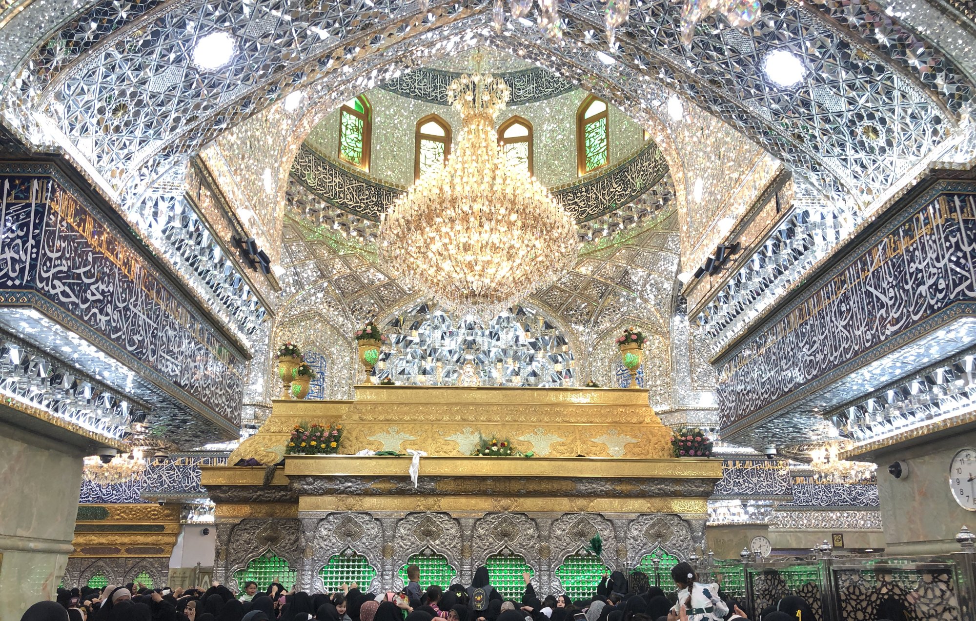 The image depicts the interior of a beautifully ornate religious site, characterized by intricate mirror work and elaborate decorations. The ceiling is adorned with shimmering tiles and a large chandelier, creating a luminous atmosphere. There is a golden structure at the center, likely a shrine or tomb, surrounded by a crowd of women wearing traditional black clothing, all appearing to be engaged in prayer or reflection. The walls and ceiling are richly decorated with calligraphy and other designs, enhancing the sacred ambiance of the space.