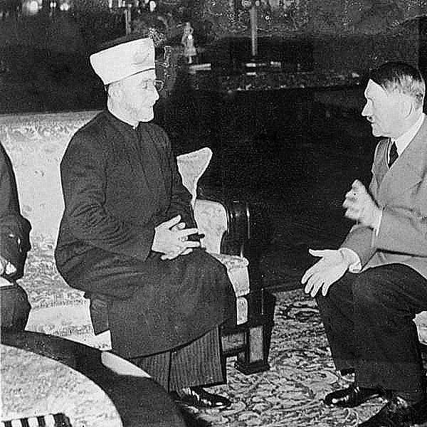 Cette image montre deux hommes assis dans une pièce. L'un d'eux porte un habit traditionnel avec un couvre-chef, tandis que l'autre est vêtu d'un uniforme militaire. Ils semblent être en conversation, gesticulant avec leurs mains. L'environnement est richement décoré, avec des meubles élégants et des éléments de décoration en arrière-plan. L'image évoque un moment de rencontre entre ces deux figures historiques.