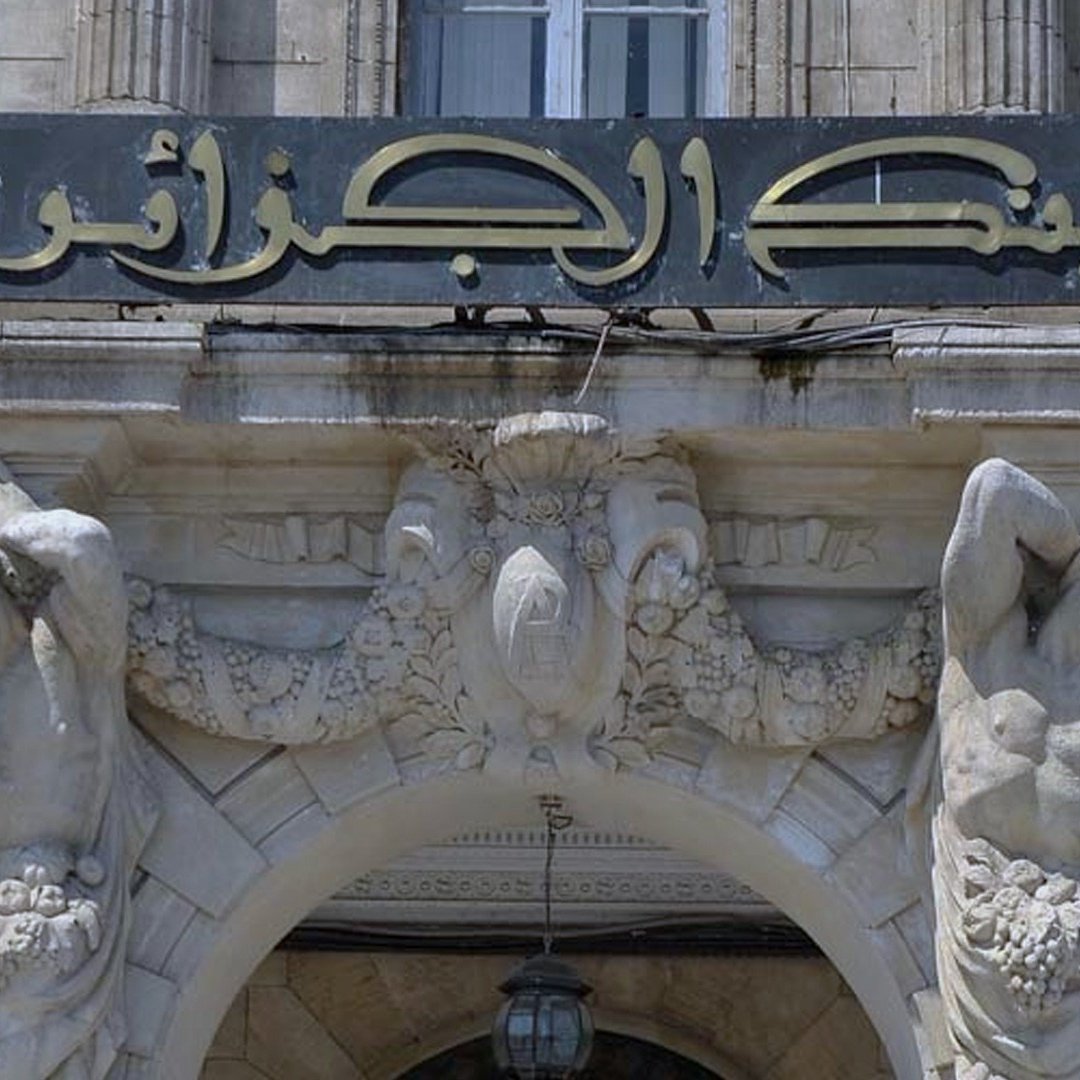 The image features a historical bank building with an ornate entrance. Above the doorway, there is a sign in Arabic that reads "بنك الجزائر" which translates to "Bank of Algeria." The architecture includes decorative elements, such as sculptures of two male figures on either side of the entrance. These figures appear to be holding or surrounded by garlands or fruit, adding to the classical decorative style of the building. The overall design is elaborate, reflecting a historical architectural style.