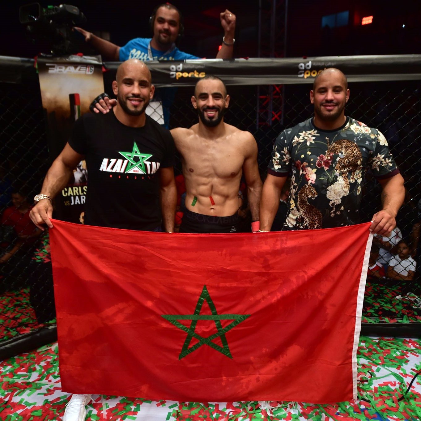 L'image montre trois hommes posant ensemble à l'intérieur d'un environnement de combat, probablement après une victoire. L'un d'eux est torse nu et affiche un large sourire, tandis que les autres sont habillés avec des t-shirts. Ils tiennent une grande drapeau marocain, mettant en avant leur fierté nationale. Le sol est couvert de confettis, ajoutant une atmosphère festive à la scène. En arrière-plan, on peut voir des spectateurs soulevant les bras en signe de célébration.
