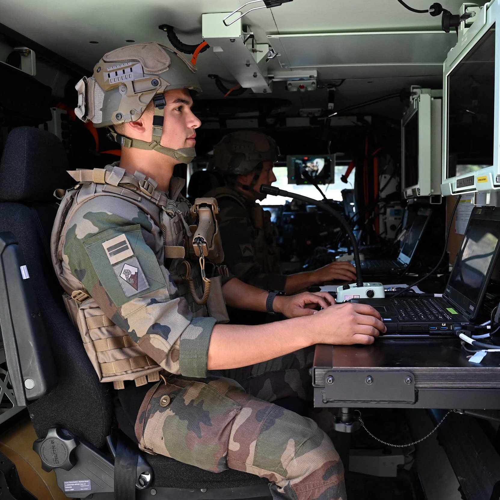 L'image montre deux soldats à l'intérieur d'un véhicule militaire. Ils portent des uniformes camouflés et des casques. L'un des soldats est assis devant un ordinateur portable, concentré sur son travail. L'intérieur du véhicule est équipé de plusieurs écrans et dispositifs technologiques. L'atmosphère semble sérieuse, indiquant une mission ou une opération en cours.