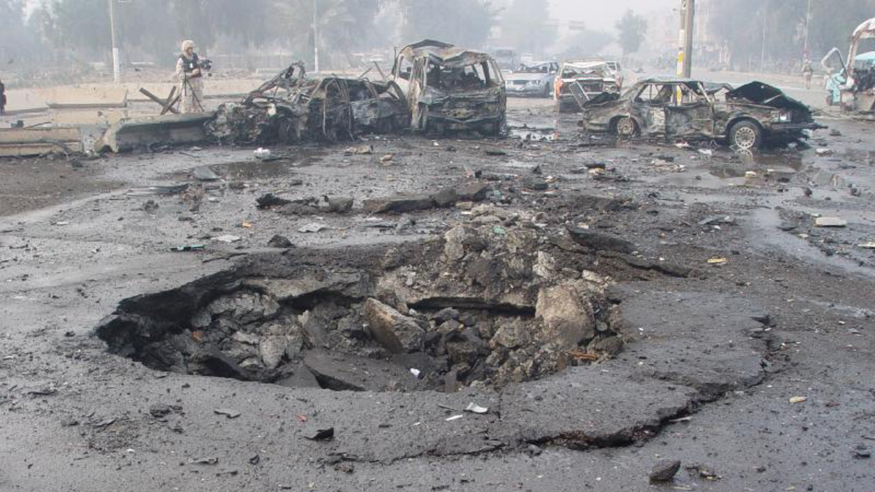 L'image montre un site de dévastation après une explosion. On peut voir plusieurs voitures endommagées et partiellement détruites, gisant sur le sol. Au centre, un cratère profond et large est présent dans le bitume, indiquant la force de l'explosion. L'environnement semble chaotique avec des débris éparpillés et une atmosphère troublée, probablement à cause de fumée ou de poussière. Cette scène évoque une situation d'urgence ou un acte de violence, reflétant des dommages importants.