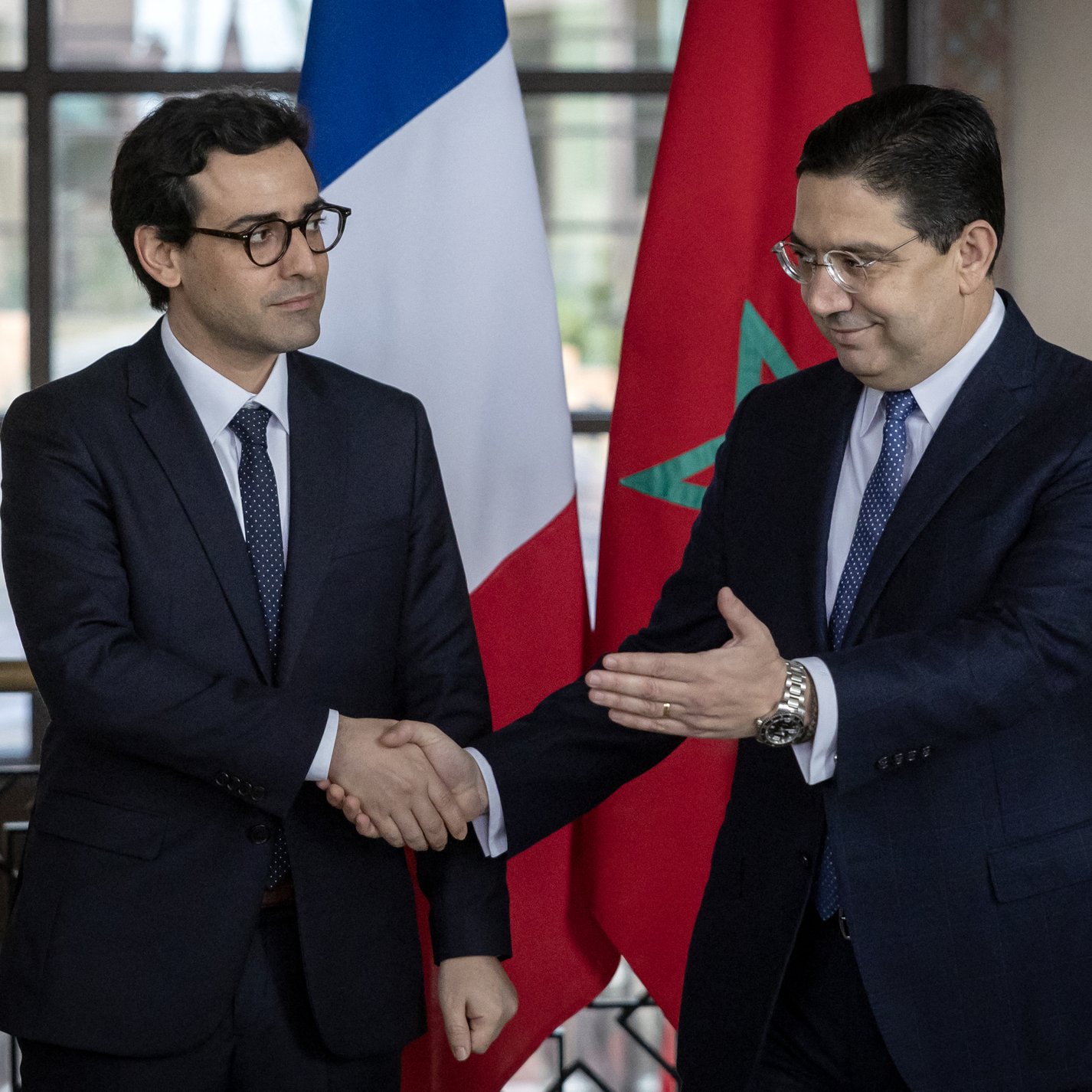 L'image montre deux hommes en costume, se serrant la main dans un cadre officiel. À l'arrière-plan, on peut voir un drapeau français à gauche et un drapeau marocain à droite. L'atmosphère semble être celle d'une rencontre diplomatique ou d'une conférence de presse, soulignant une interaction entre les deux pays.