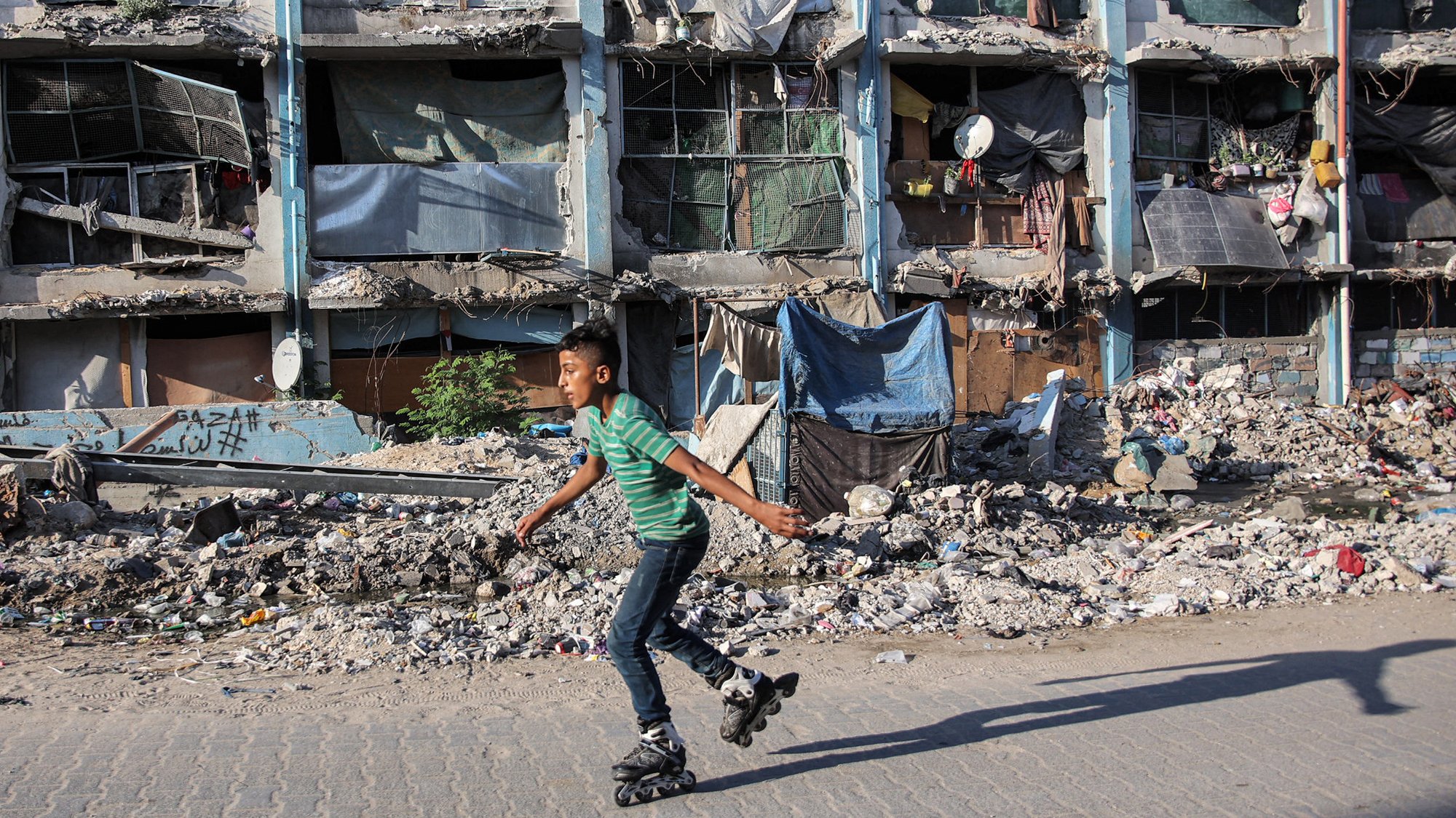 L'image montre un jeune garçon qui skates sur des rollers, se déplaçant avec agilité sur un trottoir. En arrière-plan, des bâtiments en mauvais état sont visibles, avec des fenêtres cassées et des débris éparpillés, ce qui évoque un environnement urbain dégradé. Des morceaux de tissu et d'autres objets sont accrochés aux façades, ajoutant à l'impression générale de désolation. Le contraste entre l'activité du garçon et le décor environnant souligne une certaine résilience face à la pauvreté.