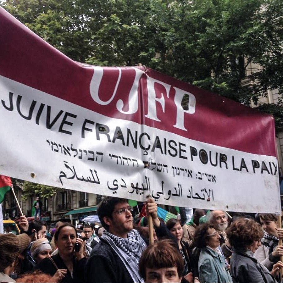 L'image montre une manifestation où des personnes tiennent une grande banderole. Cette banderole mentionne "UJFP" et affiche des messages en français, hébreu et arabe, qui semblent appeler à la paix. Le contexte suggère un rassemblement pour des causes sociales ou politiques, probablement en lien avec la paix au Moyen-Orient. En arrière-plan, on voit une foule de manifestants, des arbres et des bâtiments, ce qui indique que l'événement se déroule en milieu urbain.