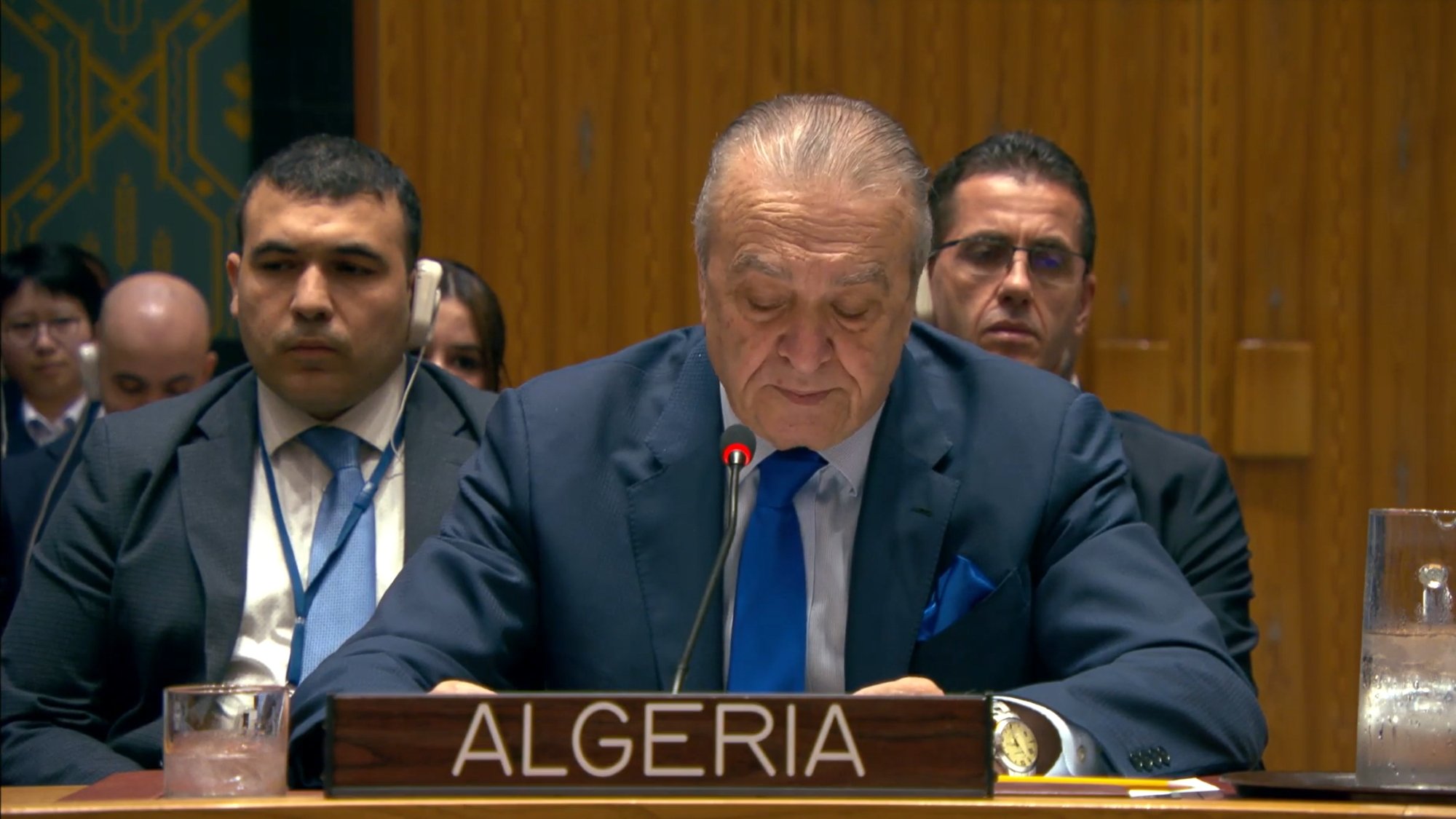 A man in a suit speaks at a podium labeled "Algeria" in a formal setting with delegates behind him.