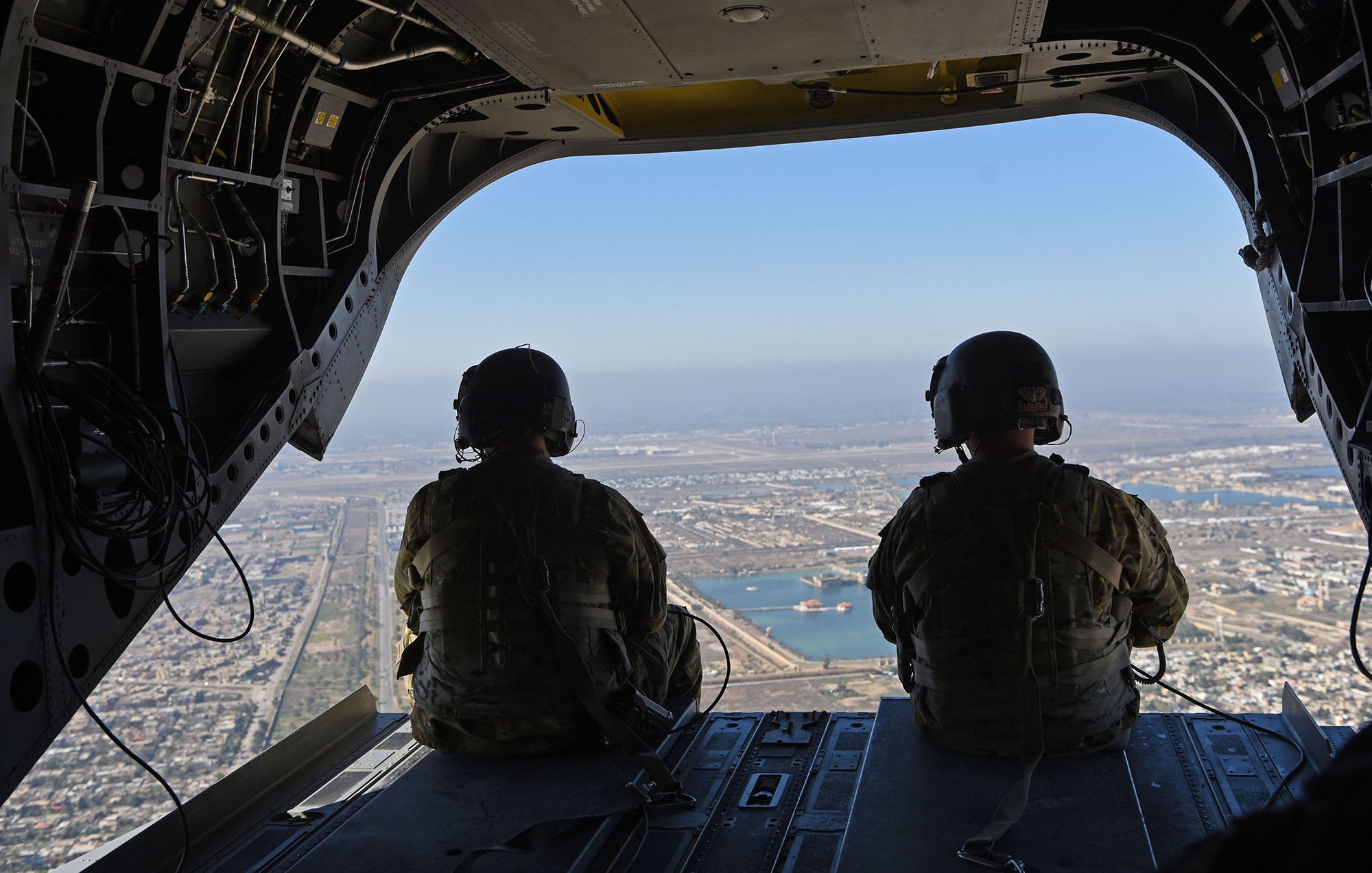 The image shows two military personnel seated inside a helicopter, looking out through the open door. They're wearing tactical gear and helmets, and the view below features a landscape with land and water. Various structures can be seen in the distance, highlighting a bustling environment beneath them. The scene conveys a sense of operation and readiness, capturing the perspective of those in flight.