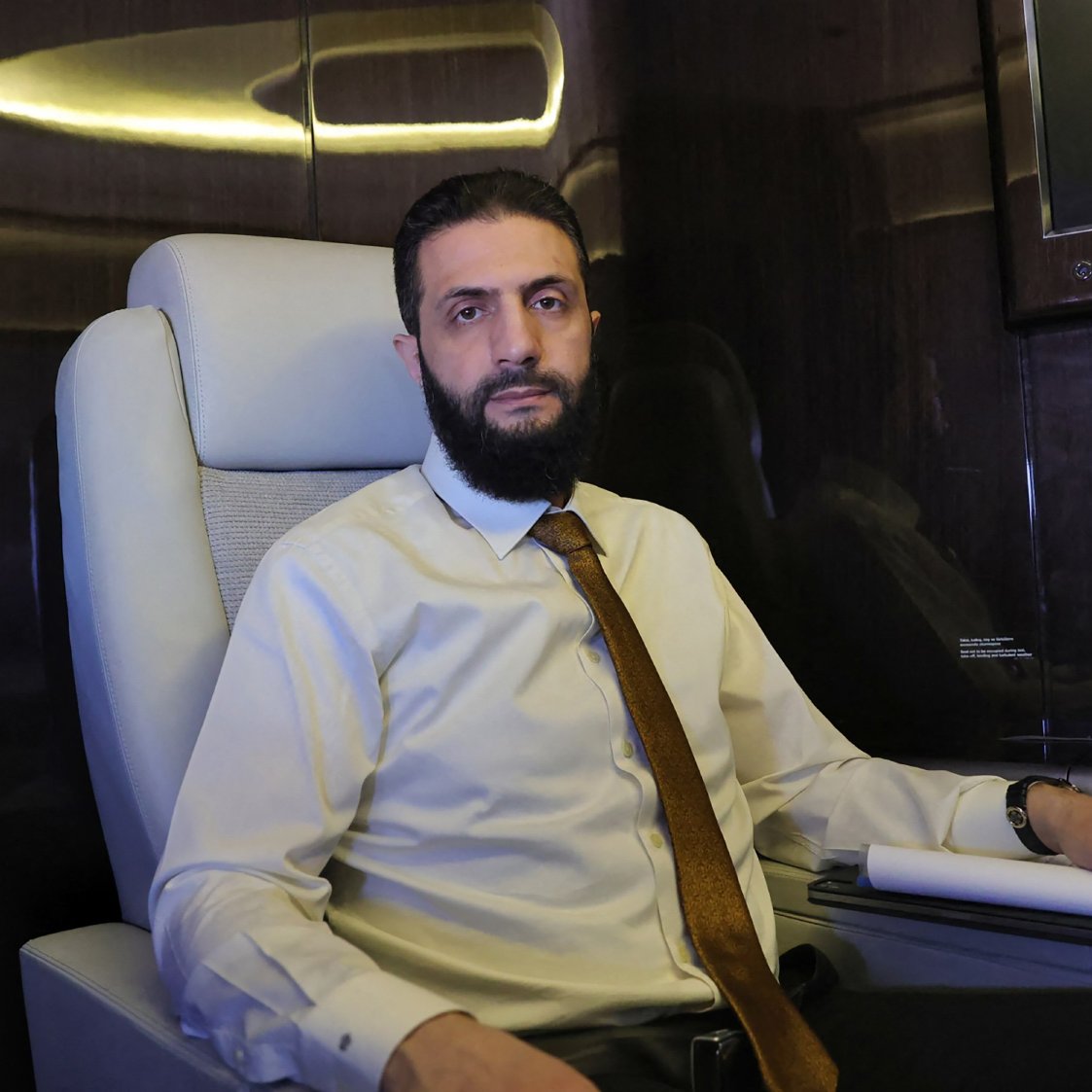 Un homme est assis dans un fauteuil confortable à l'intérieur d'un avion privé. Il porte une chemise claire et une cravate. L'environnement est élégant, avec des surfaces sombres et brillantes.