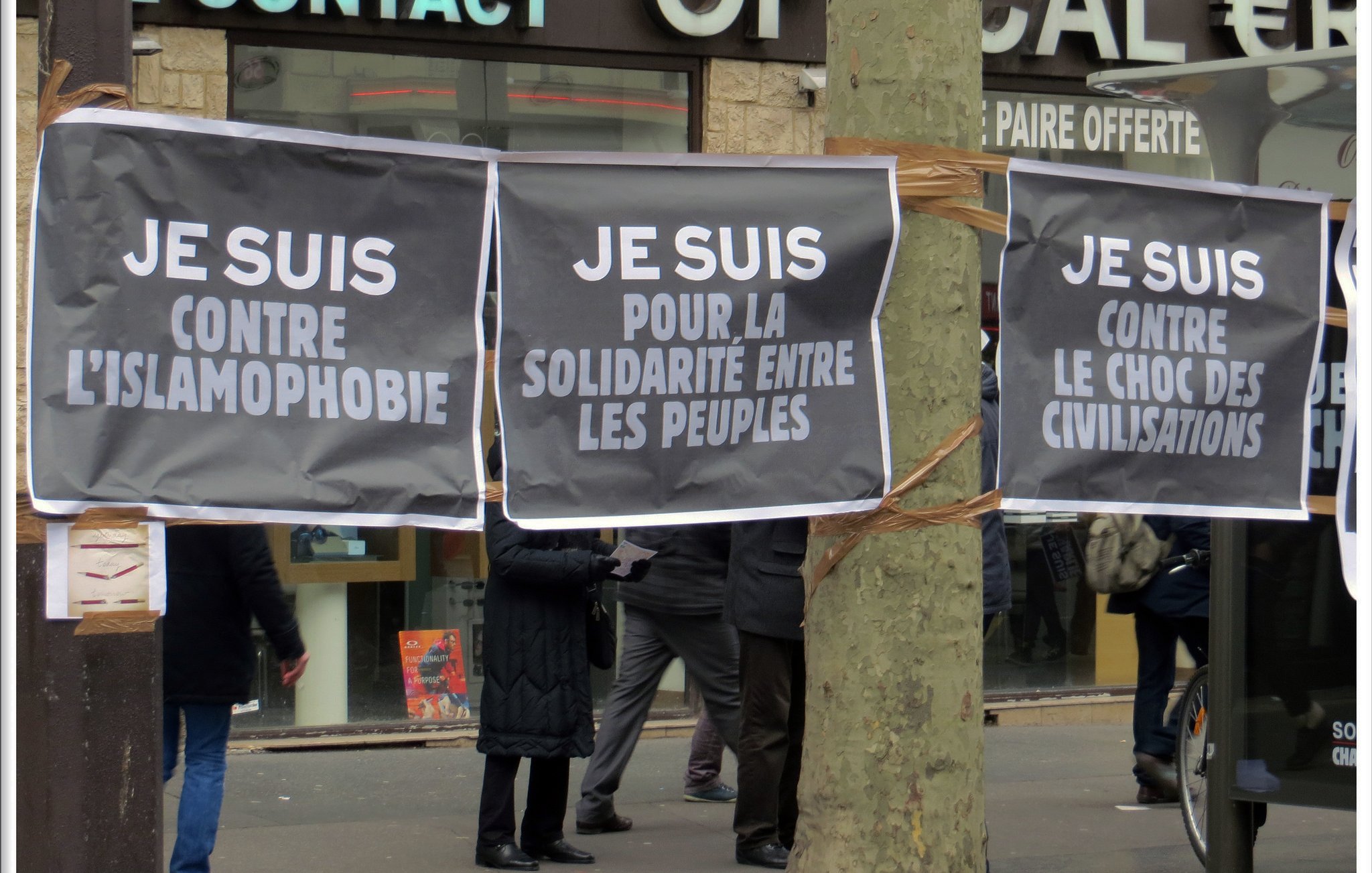 L'image montre plusieurs grandes affiches suspendues, avec des messages écrits en lettres blanches sur fond noir. Les slogans affichés expriment des positions contre l'islamophobie et en faveur de la solidarité entre les peuples. En arrière-plan, on peut apercevoir des personnes qui semblent participer à un rassemblement ou une manifestation. L'ambiance suggère une mobilisation citoyenne pour des causes sociales et politiques.