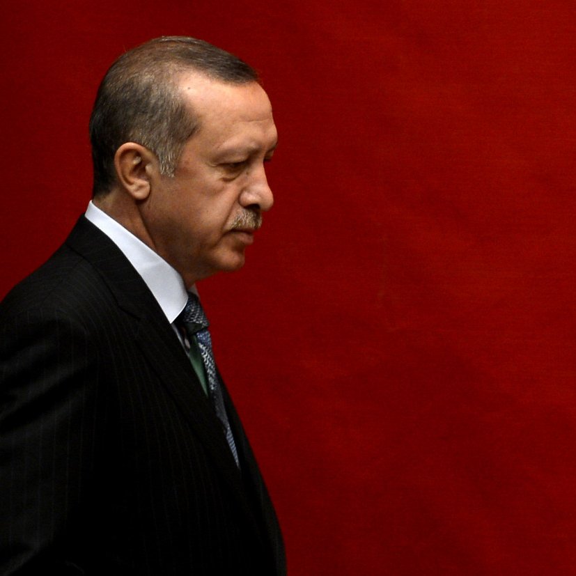 L'image montre un homme en costume sombre, marchant sur le côté, avec une expression sérieuse. Le fond est rouge uni, créant un contraste marqué avec sa tenue. L'éclairage met en avant son profil et accentue les détails de son visage.