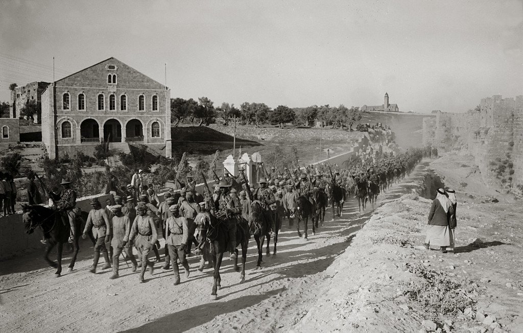 L'image montre une troupe de soldats à cheval marchant en formation le long d'un chemin. En arrière-plan, on peut voir des bâtiments en pierre, dont une grande structure avec des fenêtres. Le sol est poussiéreux, et on remarque également quelques personnes à pied, dont une femme en robe. L'atmosphère semble être celle d'une époque historique, probablement d'un conflit ou d'une quelconque mobilisation militaire. Les soldats portent des uniformes similaires et semblent déterminés dans leur avancée.