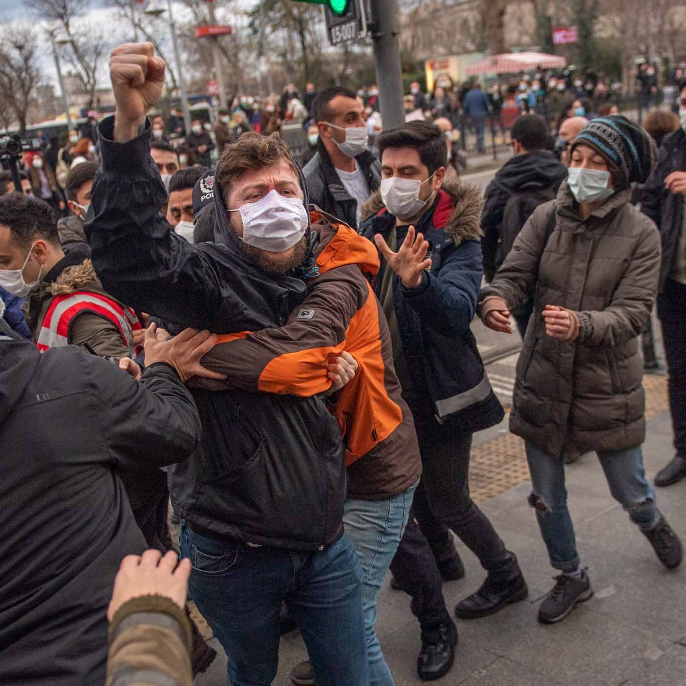 در تصویر، گروهی از جوانان در حال برگزاری یک تجمع هستند. برخی از افراد ماسک به صورت دارند و به نظر می‌رسد که در حال ابراز نظرات یا اعتراضات خود هستند. یکی از افراد به وضوح در حال فریاد زدن و دستش را به هوا بلند کرده است، در حالی که دیگران او را نگهداشته‌اند. به نظر می‌رسد وضعیت هیجانی است و برخی از افراد در تلاش برای برقراری نظم یا جلوگیری از درگیری هستند. پشت زمینه نیز شهری را نشان می‌دهد که با تابلوهای ترافیکی و ساختمان‌ها مشخص شده است.