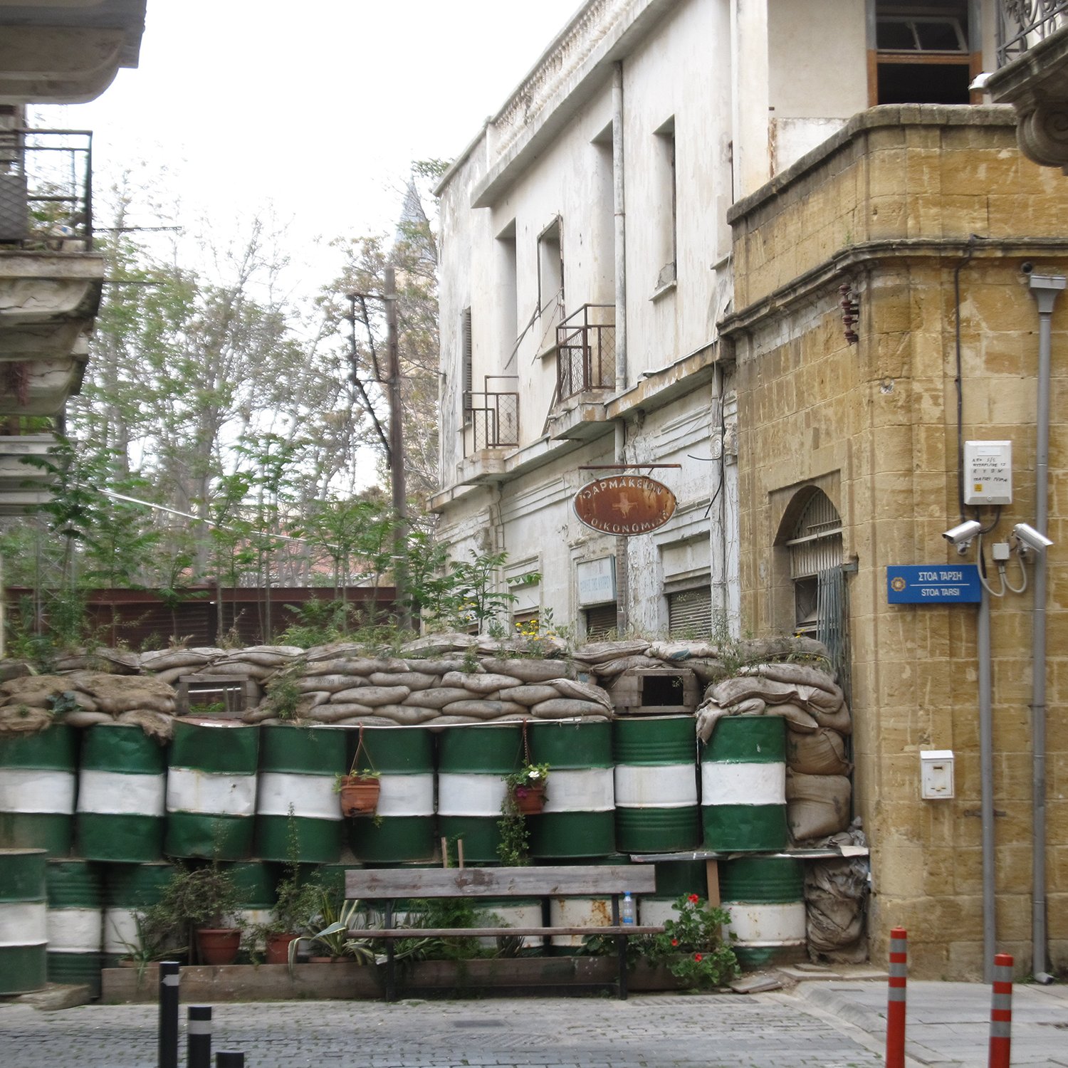 L'image montre une rue étroite avec un mur constitué de sacs de sable et de fûts en plastique, probablement utilisés comme une barrière de protection. Au fond, il y a des bâtiments anciens avec des balcons, et des signes de végétation qui poussent sur le mur. L'atmosphère de la scène semble être à la fois urbaine et quelque peu abandonnée, avec un mélange de vie naturelle et d'éléments de construction.