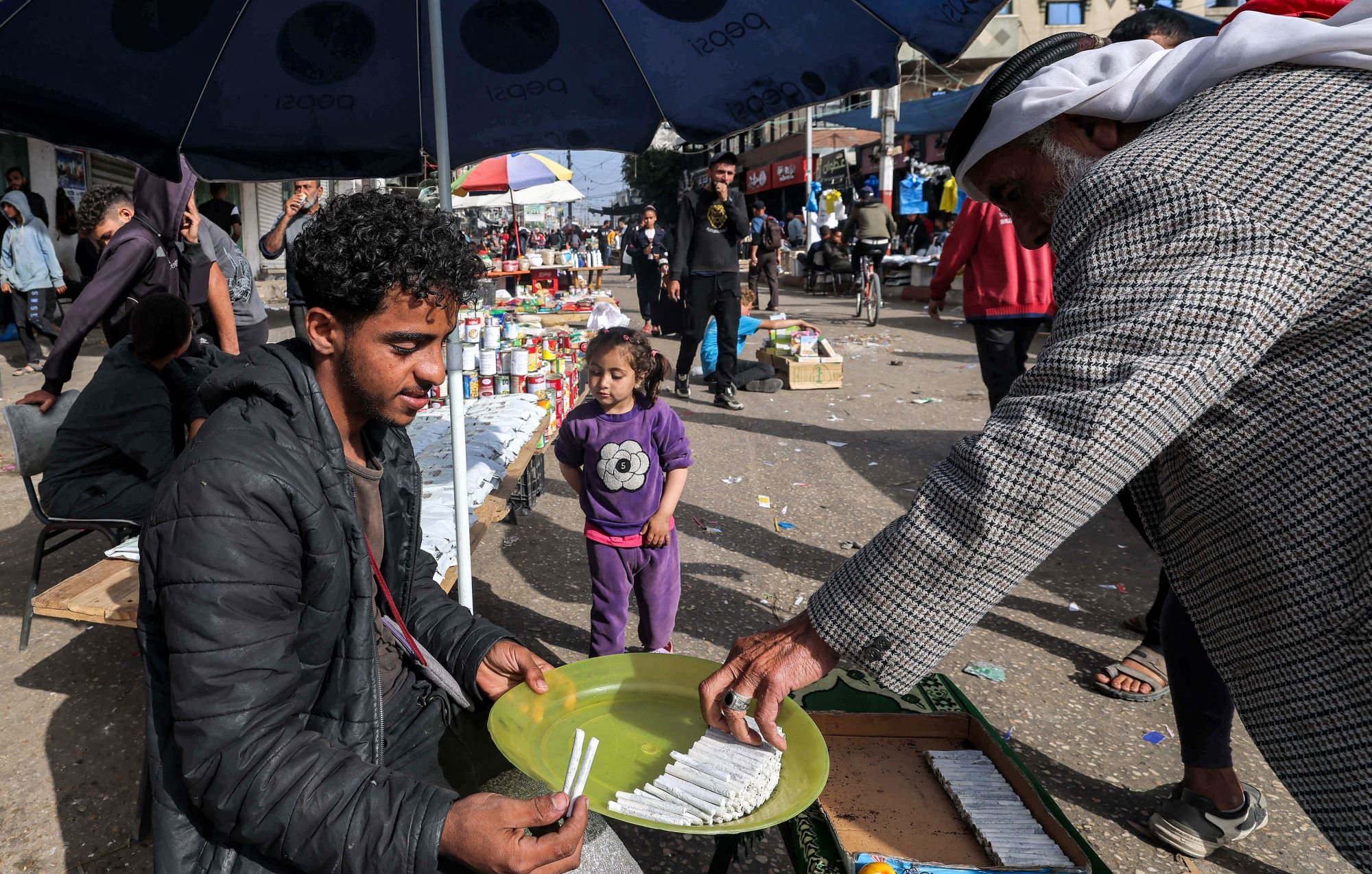 در تصویر، یک بازار محلی به چشم می‌خورد. در وسط تصویر، مرد جوانی نشسته و در حال فروش کالاهایی است. او یک بشقاب سبز رنگ را در دست دارد که در آن چند قلم وجود دارد. به نظر می‌رسد که او به مشتری‌اش توجه دارد که در حال انتخاب کالاهاست. در کنار آن، یک دختر بچه نیز در حال بازی کرده و به صحنه نگاه می‌کند. در پس‌زمینه، افراد دیگری در حال خرید و گشت و گذار هستند و جوی شلوغ و پرجنب‌وجوش به چشم می‌خورد. سایه چترهایی که بالای سر برخی افراد است، فضایی دلپذیر ایجاد کرده است.
