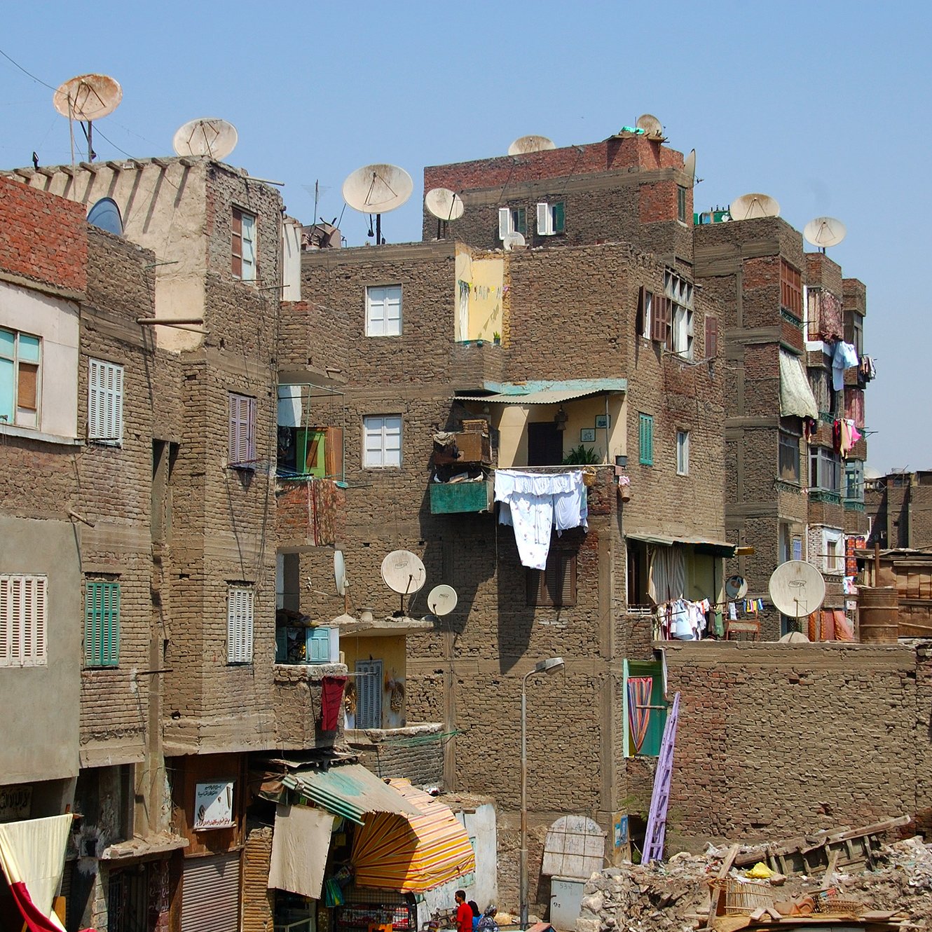 L'image montre un paysage urbain d'un quartier populaire. On peut voir des bâtiments en briques de terre, souvent de plusieurs étages, avec des façades colorées. De nombreuses antennes parabolique sont installées sur les toits. Les vêtements sont accrochés à l'extérieur pour sécher. L'atmosphère semble animée, avec des signes de vie quotidienne. Le ciel est dégagé, ce qui donne une impression de chaleur.