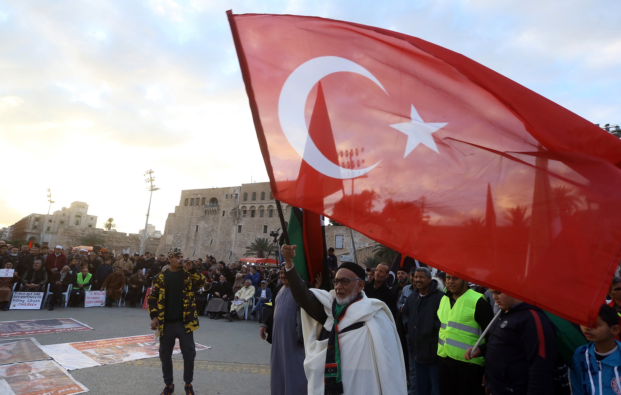 L'image montre une scène de rassemblement ou de manifestation en plein air. Au premier plan, un homme âgé tient fièrement un grand drapeau turc, qui est rouge avec une étoile et un croissant blancs. On peut apercevoir une foule de personnes en arrière-plan, certaines portant des vêtements de sécurité fluorescents. La scène se déroule peut-être dans un lieu historique, avec un bâtiment ancien visible derrière la foule. L'ambiance semble dynamique et engagée.