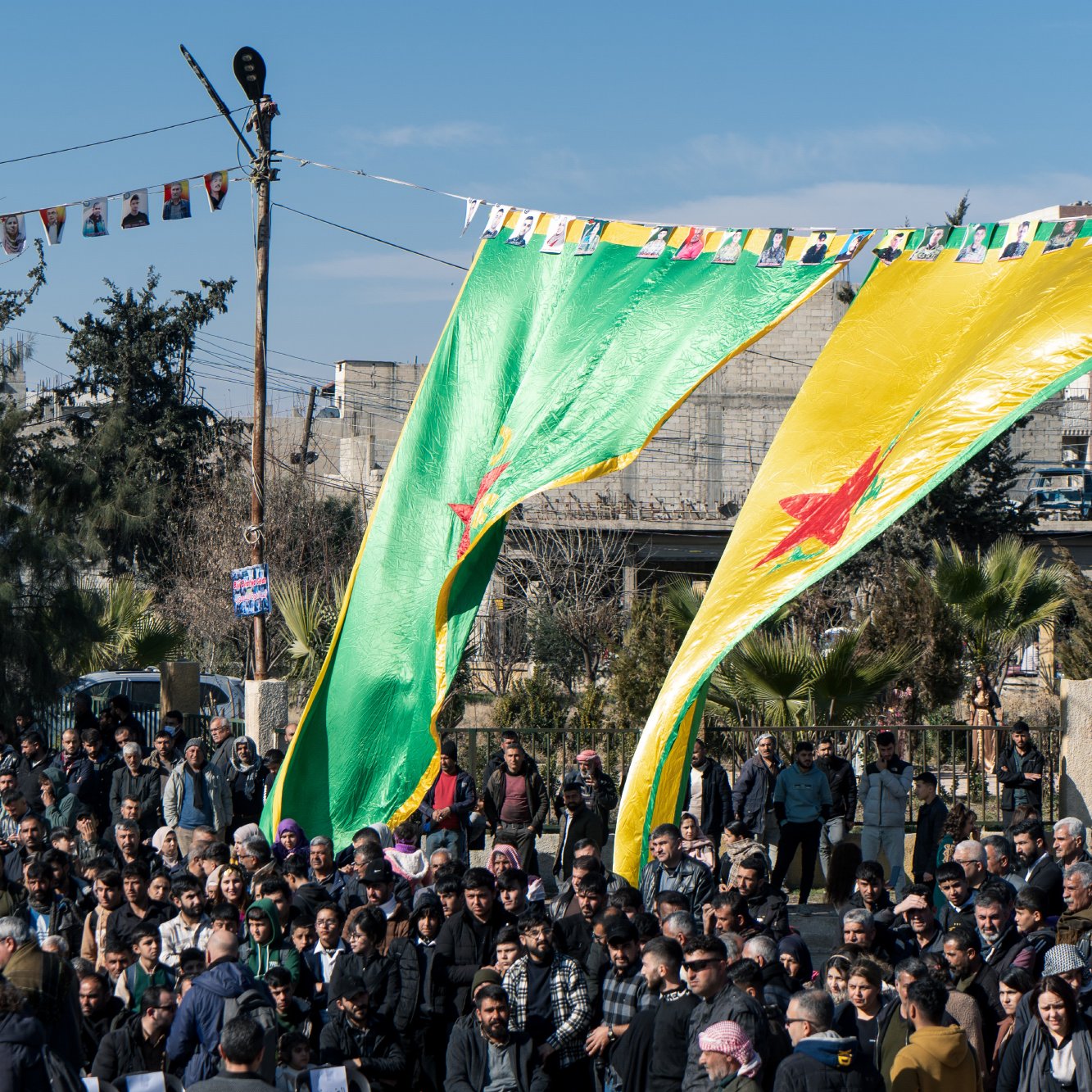 L'image montre une foule rassemblée sous de grandes drapeaux verts et jaunes, portant une étoile rouge. En arrière-plan, des bâtiments et des arbres sont visibles. Des banderoles sont accrochées au-dessus des personnes, indiquant une célébration ou un rassemblement communautaire. L'atmosphère semble vibrante et engagée, avec un grand nombre de participants.
