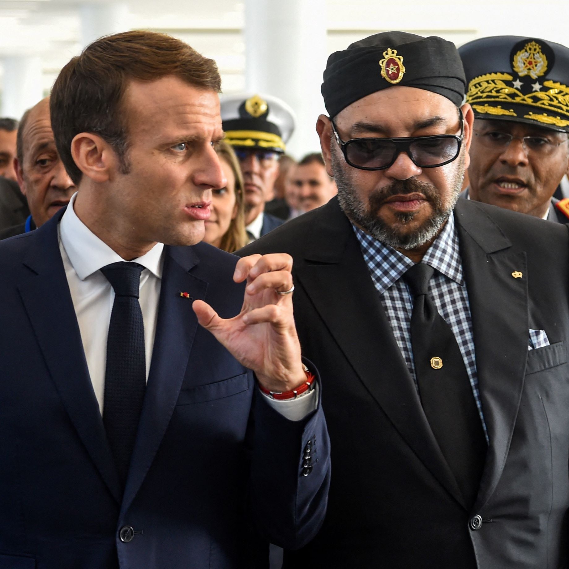 الصورة تظهر شخصين يتحدثان معاً في حدث رسمي. الشخص على اليمين يرتدي نظارات شمسية وملابس داكنة مع ربطات عنق، بينما الشخص على اليسار يرتدي بدلة رسمية ويتحدث بحركة يد معبّرة. خلفهما يوجد عدد من الأشخاص، مما يشير إلى أنهم في تجمع أو مؤتمر. الجو العام يبدو رسمياً وجاداً.