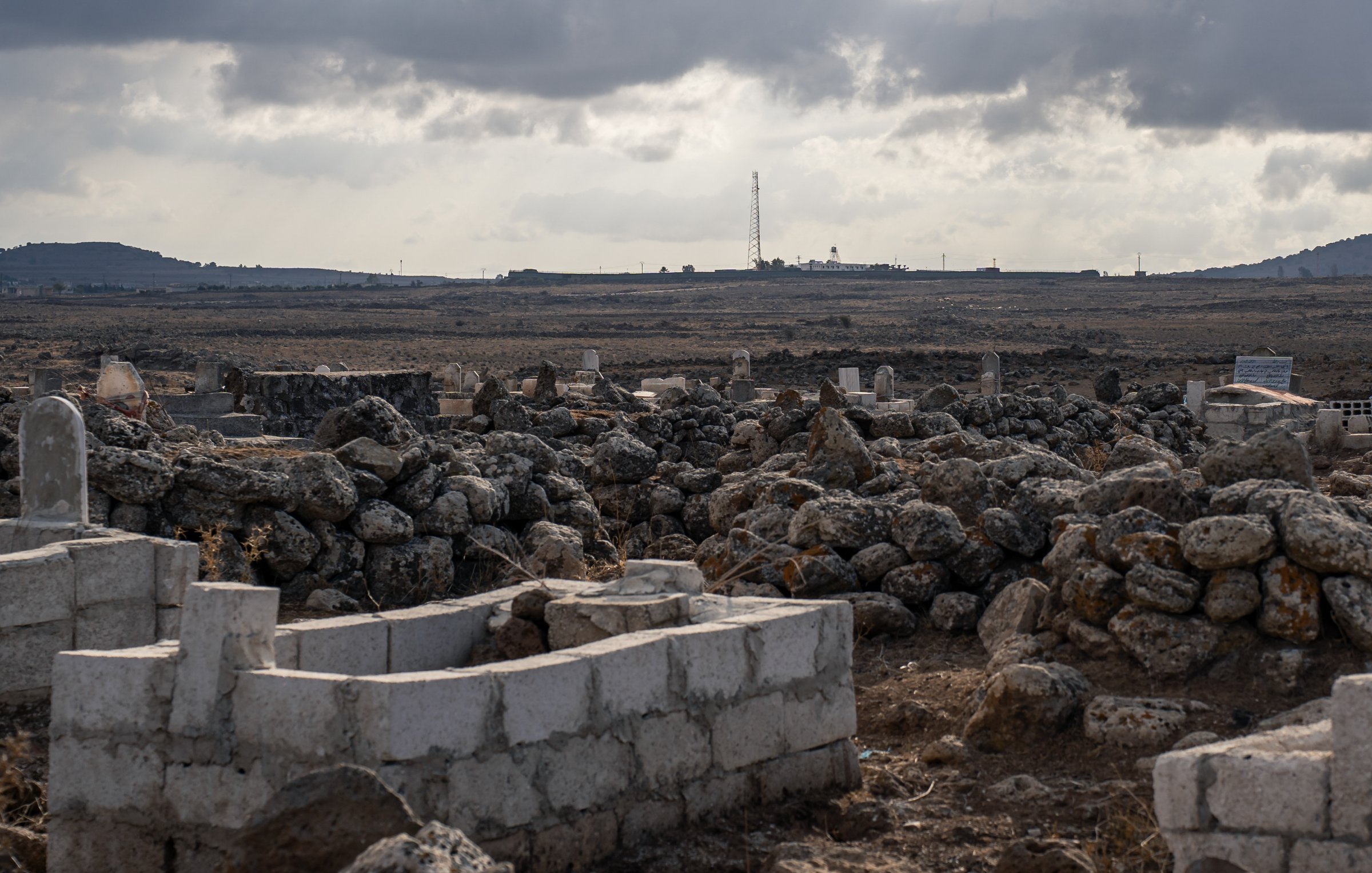 Un paisaje árido con tumbas y rocas, bajo un cielo nublado y distante, se ve una torre.