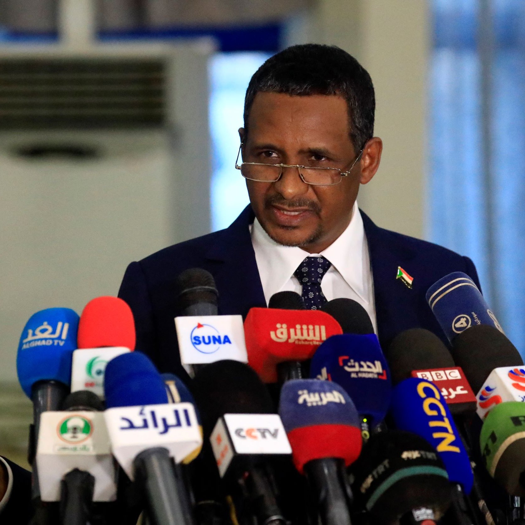 L'image montre un homme en costume, se tenant devant un grand regroupement de microphones provenant de différentes chaînes de télévision et médias. Il semble être en train de faire une déclaration ou un discours, avec des gestes expressifs. L'environnement est formel, suggérant une conférence de presse ou un événement politique.