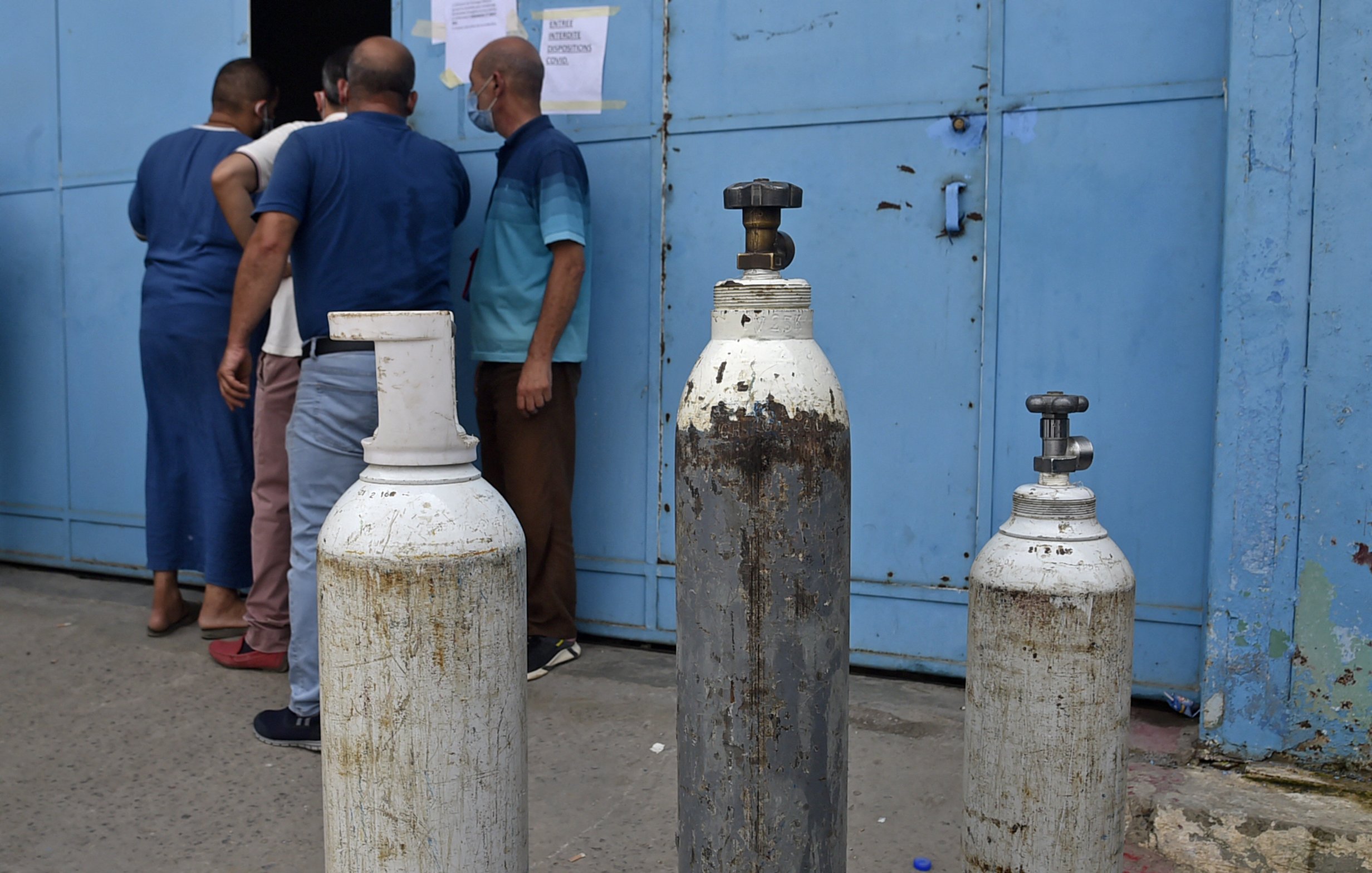 تظهر الصورة مجموعة من أسطوانات الغاز التي تُستخدم عادةً في المستشفيات أو للأغراض الصناعية. توجد ثلاث أسطوانات بأحجام متفاوتة، وتبدو واحدة منها قديمة ومُهملة. في الخلفية، يمكن رؤية بعض الأشخاص يقفون بالقرب من باب مبلوطة باللون الأزرق، مما يشير إلى موقع ربما يكون مركزًا لتوزيع الغاز أو خدمة طبية. تبدو الأجواء حيّة مع تواجد الأشخاص، مما يُبرز أهمية تلك الأسطوانات في الحياة اليومية.