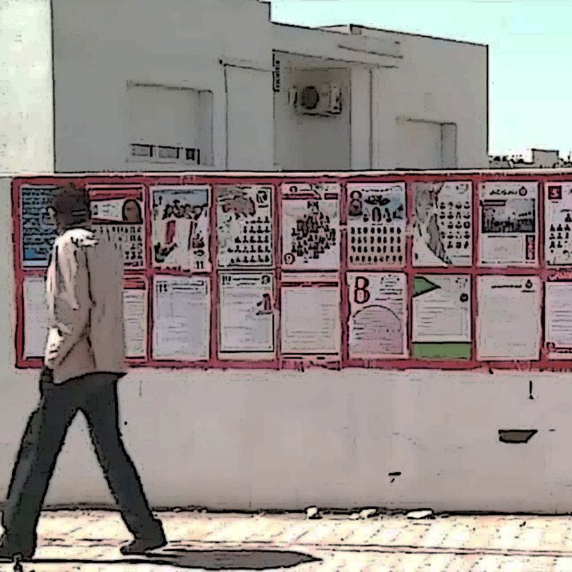 L'image montre un mur recouvert de plusieurs affiches et panneaux d'information. Ces affiches semblent contenir des annonces, des avis ou des informations diverses, affichées de manière colorée. Un homme marche devant ce mur, ce qui donne une idée de l'échelle des affiches et de l'environnement. Le paysage est lumineux, suggérant une journée ensoleillée.