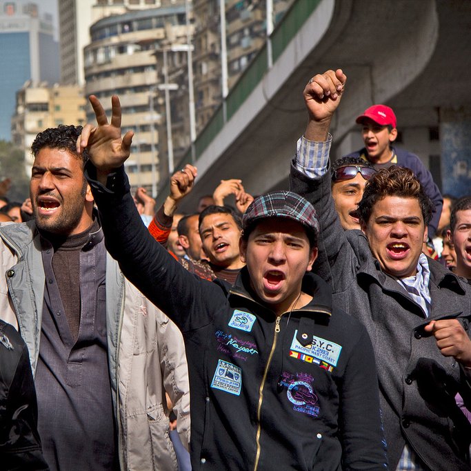 L'image montre un groupe de jeunes manifestants dans une rue animée. Ils expriment des émotions fortes, avec des visages déterminés et des gestes de protestation. Certains lèvent les bras et serrent les poings, tandis que d'autres crient. L'arrière-plan présente des bâtiments urbains, et l'atmosphère générale semble être celle d'une mobilisation collective. Les personnes sont habillées de manière variée, reflétant un mélange de styles.