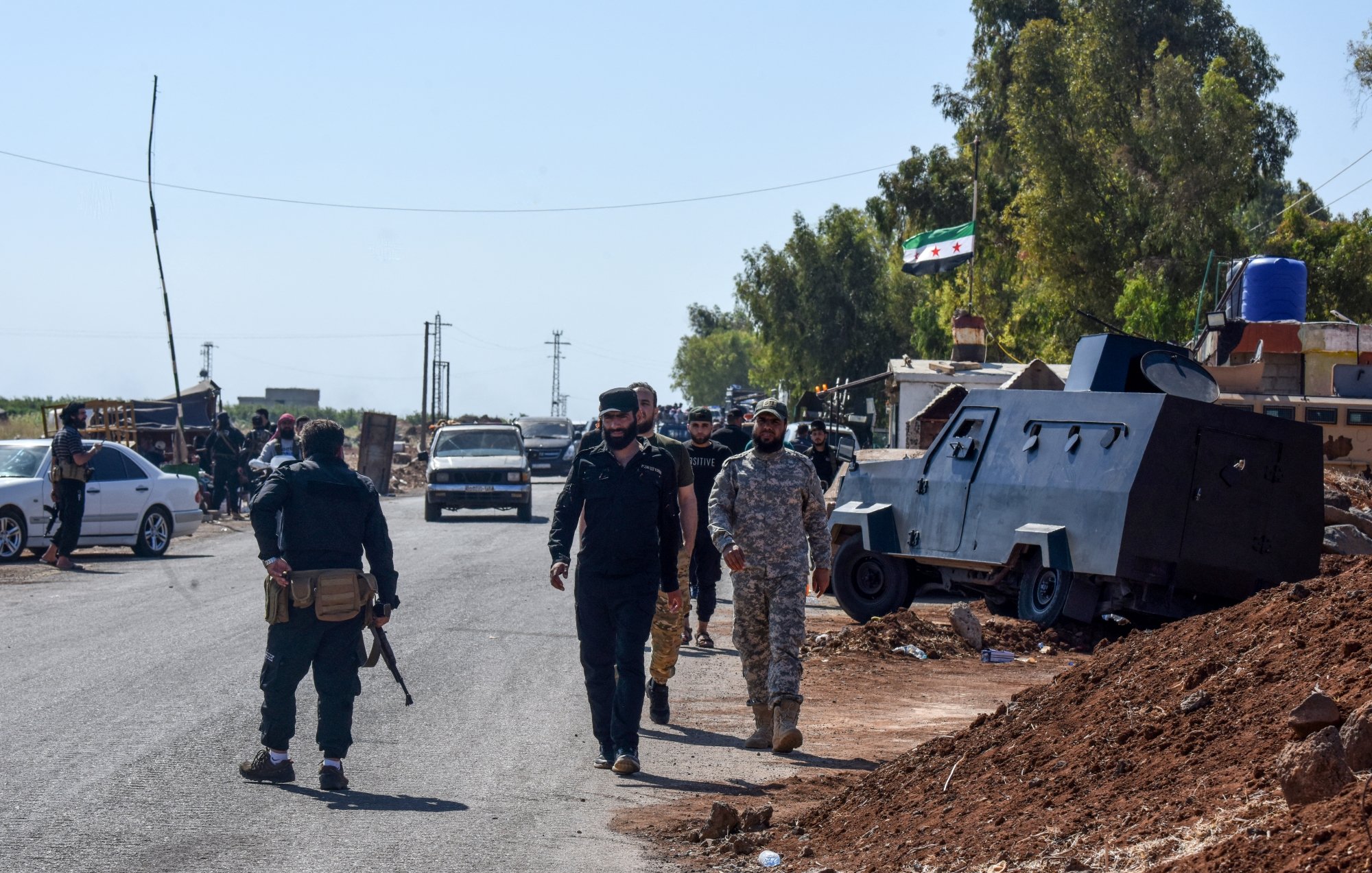 تصویر، یک خیابان با خودروها و کامیون‌ها است. چند نفر با لباس نظامی در حال حرکت هستند. دیوارها و درختان اطراف هستند.